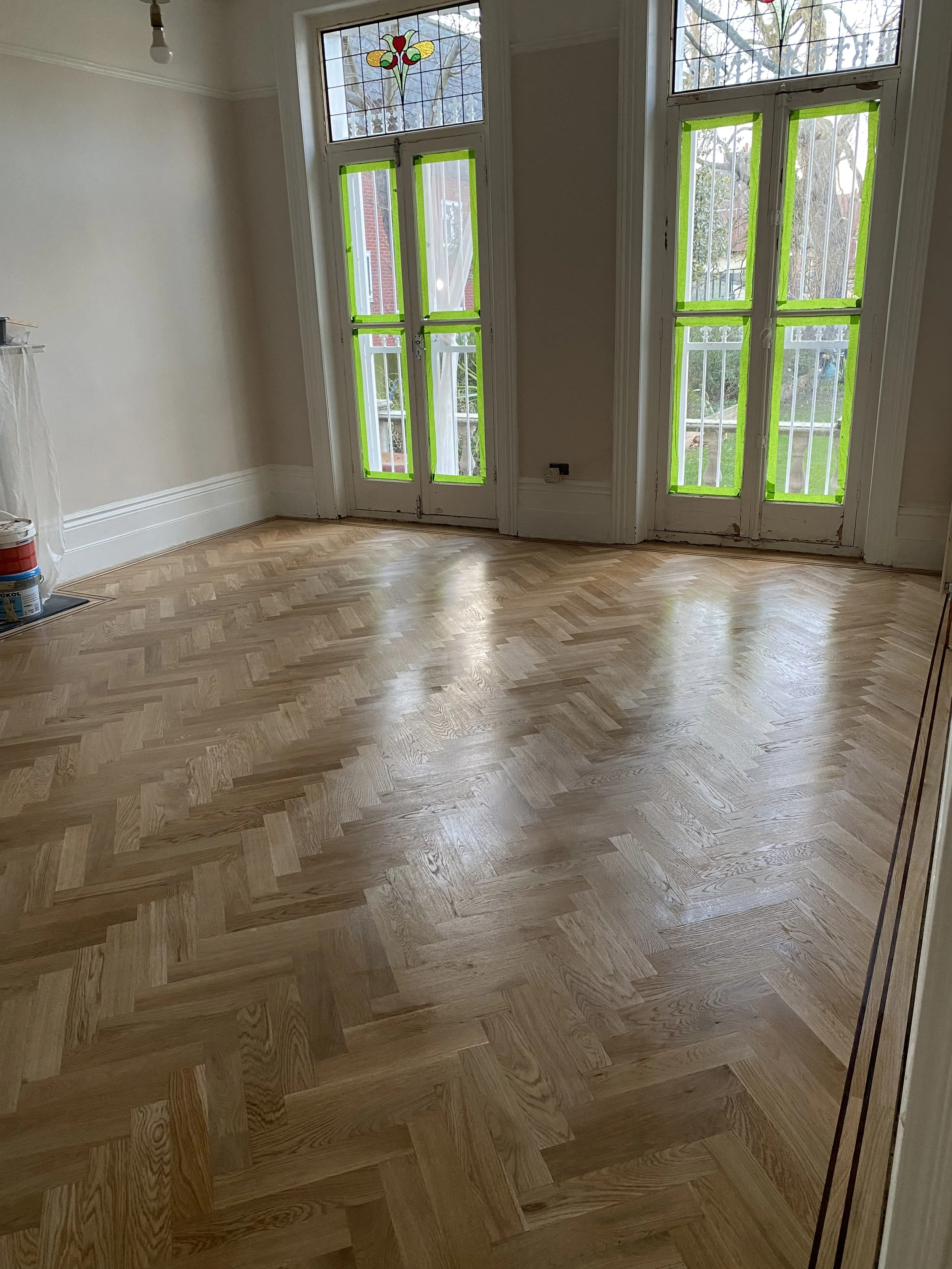 Empty room with newly installed wooden parquet floor, stained glass window, and French doors with green painter's tape on the panes.