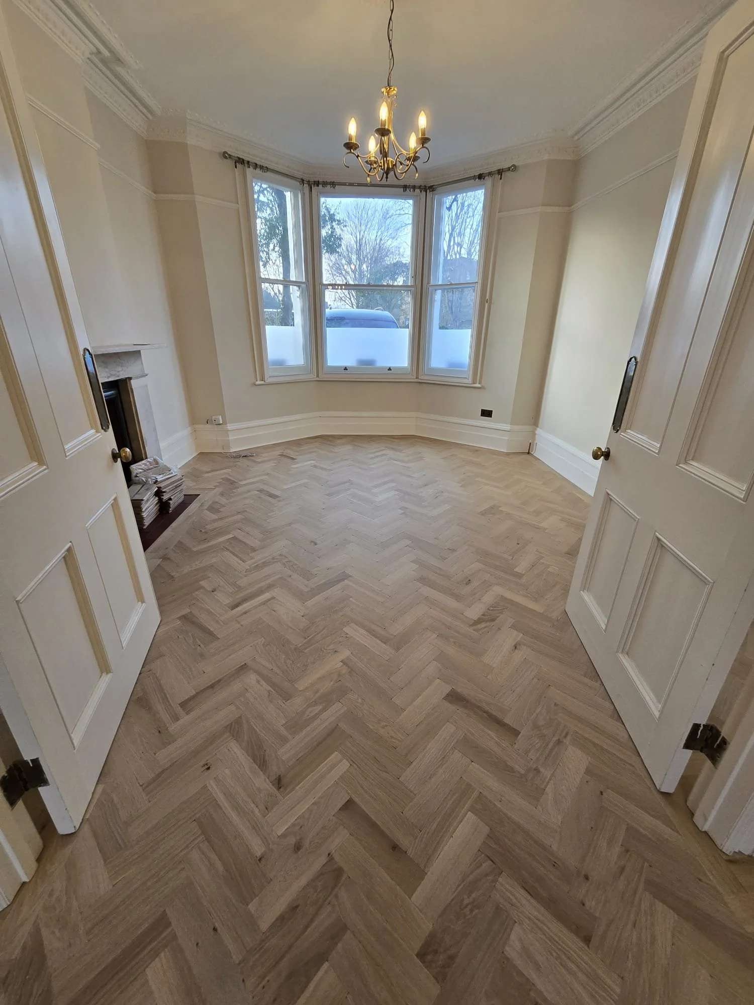 Empty room with large bay window, parquet wood floor in herringbone pattern, chandelier, and a fireplace