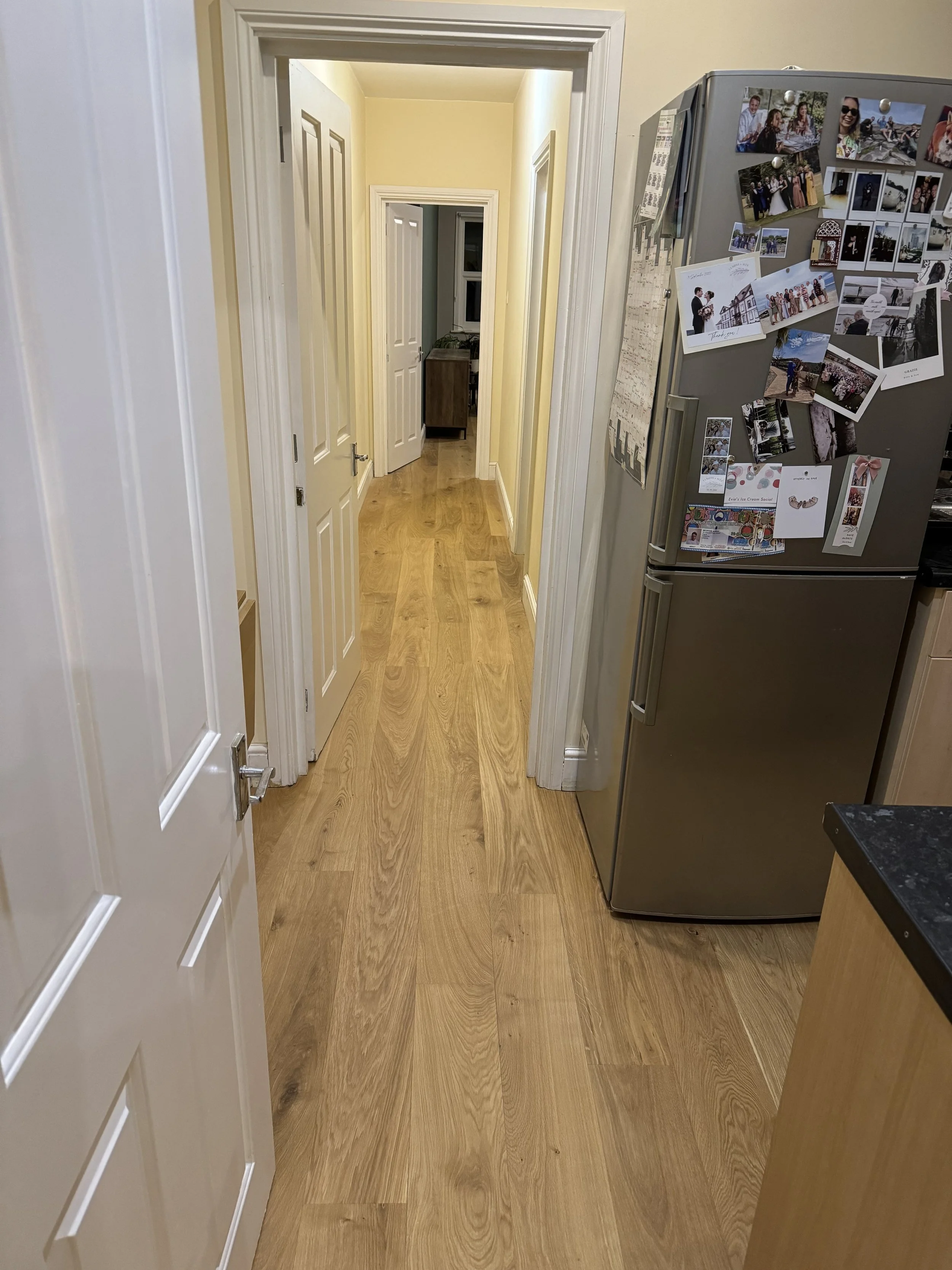A view of a hallway inside a house with wooden flooring, white doors, and a yellow-painted wall. A refrigerator covered with photos and notes is on the right side.