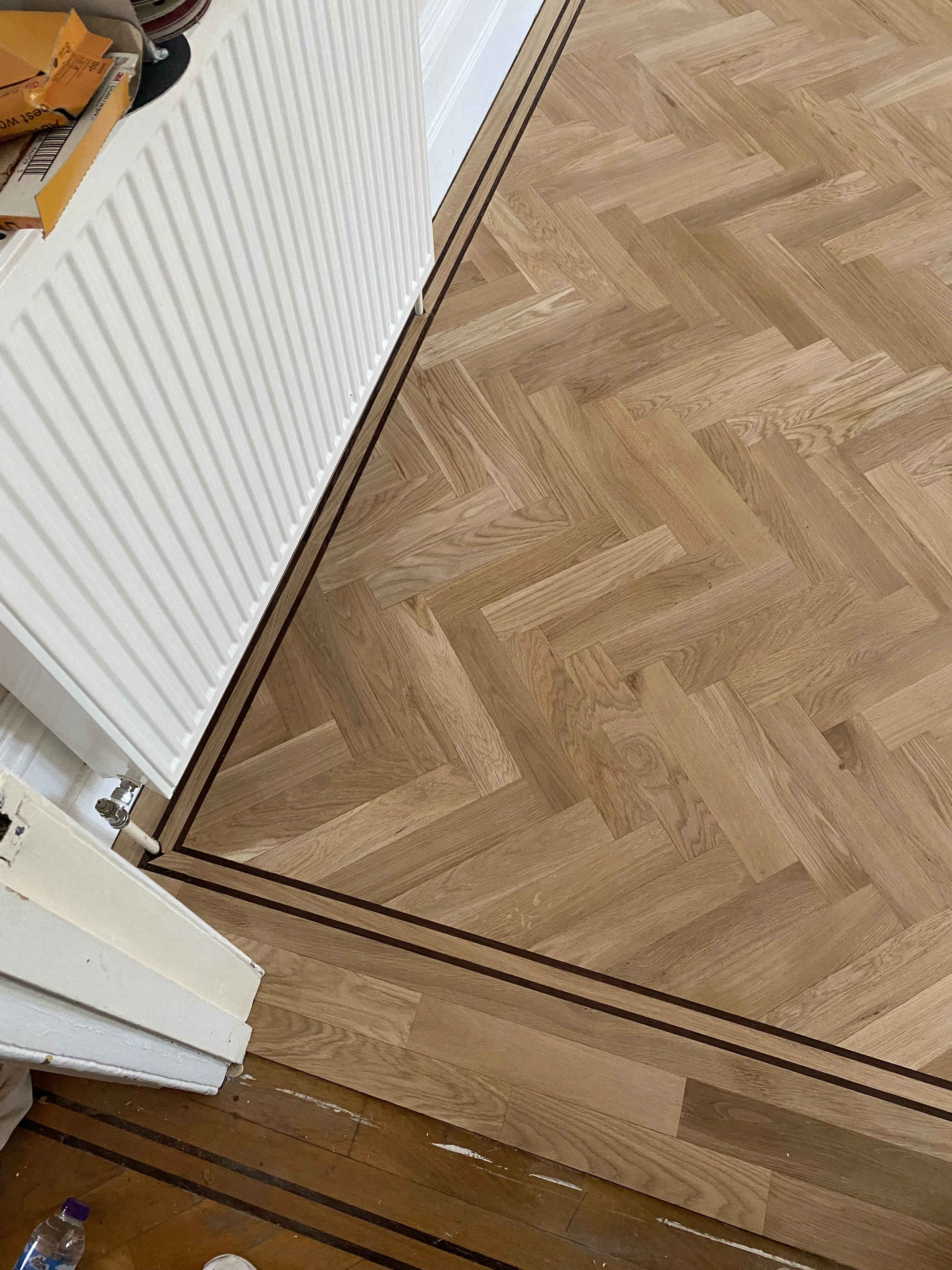 Close-up of a corner of a room showing a wooden floor with a herringbone pattern and a white radiator mounted on the wall. Part of the wall and a box of items are also visible.