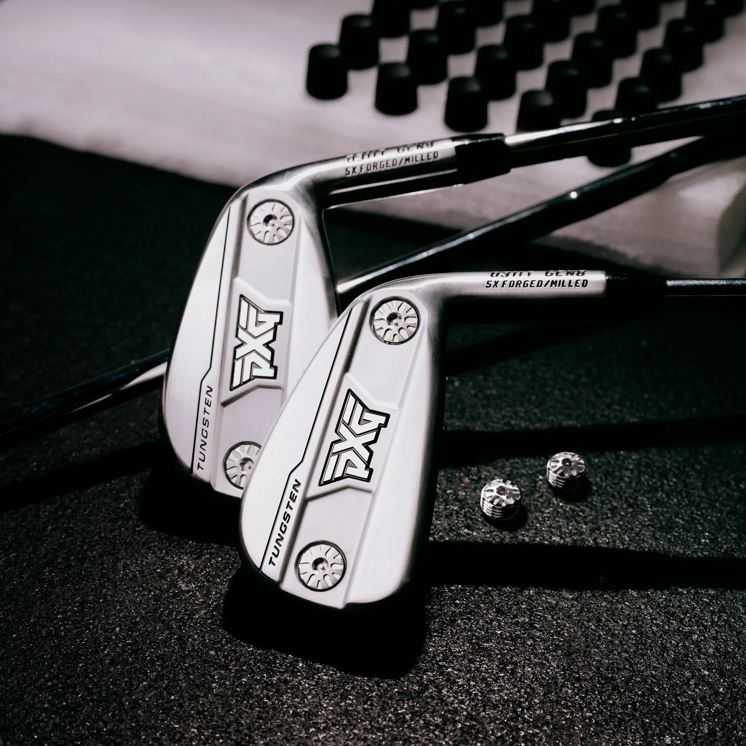 Close-up of two golf club wedges with black grips on a black textured surface, with a few silver golf club weights next to them.