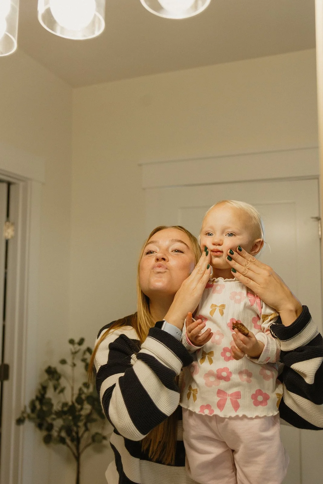 A woman with long red hair is holding a young girl with blonde hair in front of a mirror. The woman is making a puckered face while the girl is touching her face and holding a cookie, with a neutral expression. The woman is wearing a black and white striped sweater, and the girl is in a floral shirt and light pants. A plant with dark green leaves is visible in the background, and the ceiling has a modern light fixture.