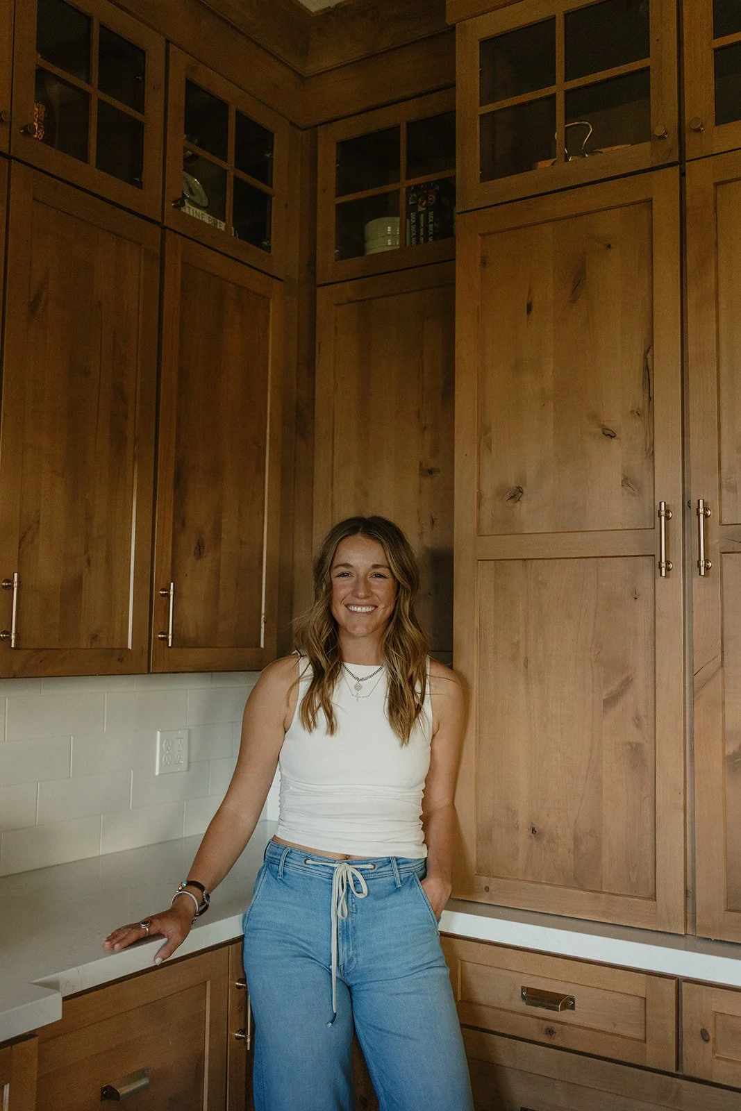 A woman with long wavy hair wearing a white sleeveless top and blue drawstring pants standing in a kitchen with wooden cabinets and a white countertop, smiling at the camera.