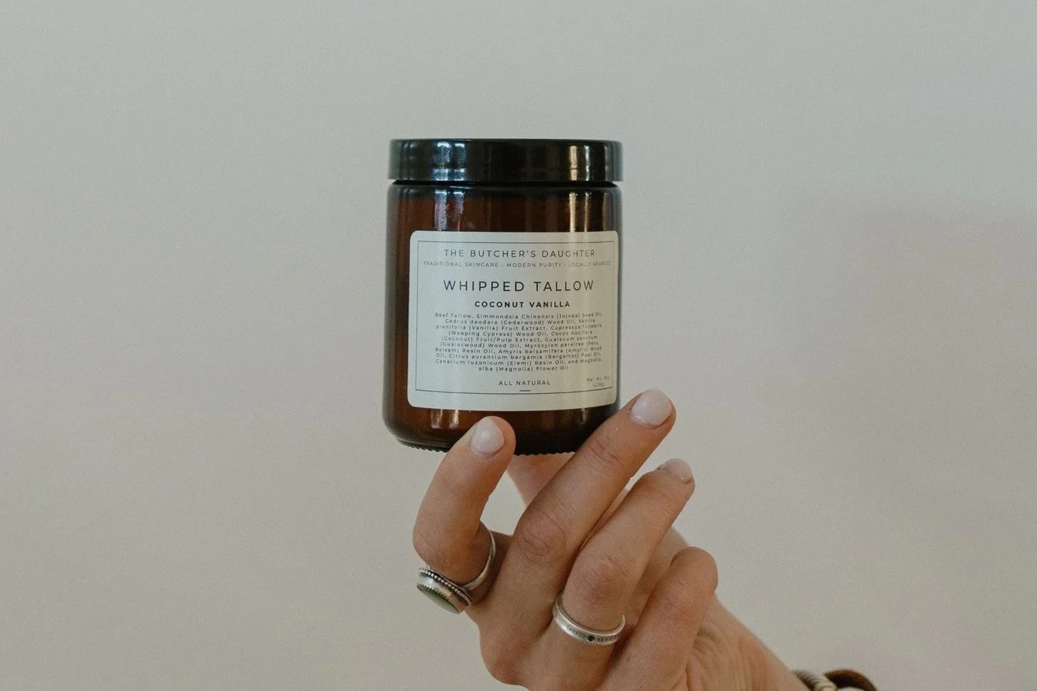 A person holding a jar of whipped tallow with coconut vanilla scent against a plain, light-colored background.