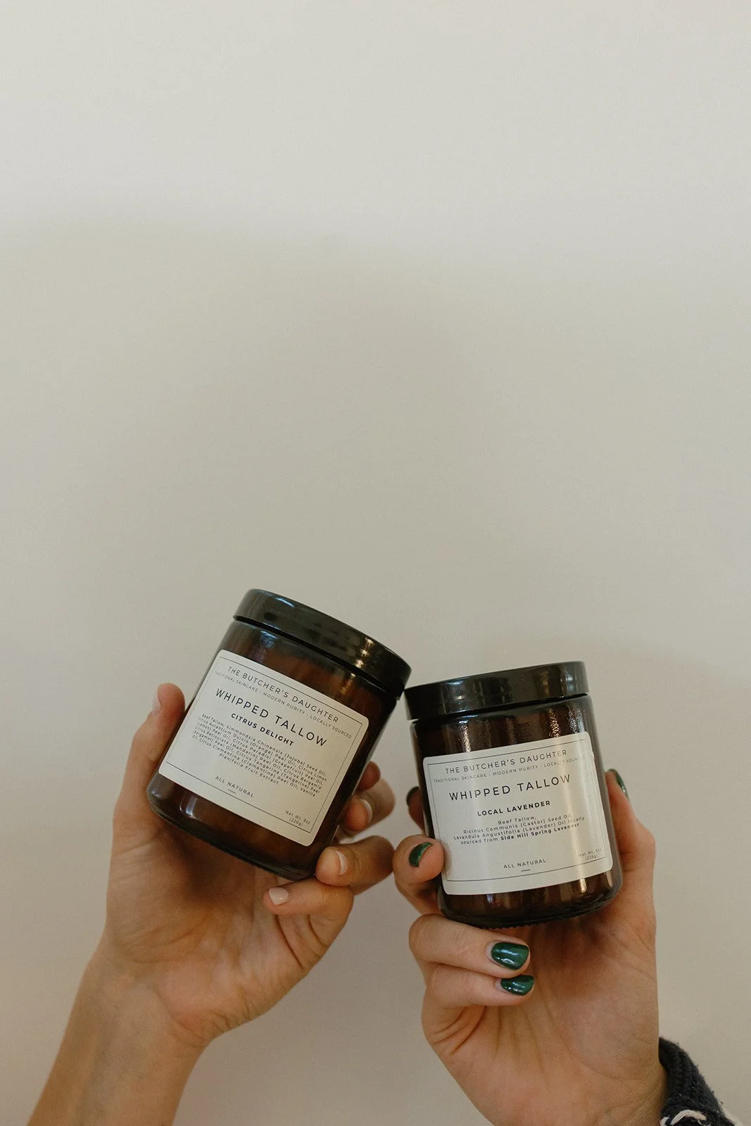 Two hands holding brown jars of whipped tallow labeled "The Butcher's Daughter" against a white background.