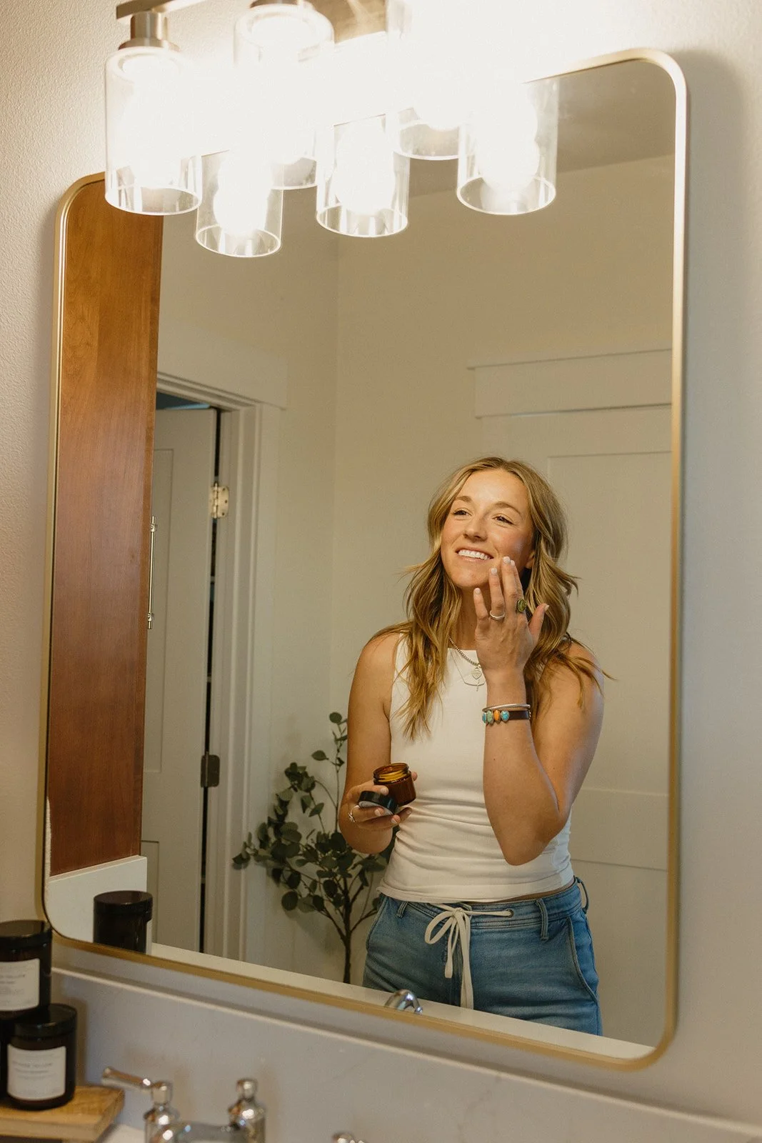 A woman taking a mirror selfie in a bathroom, smiling while applying lotion to her face, with skincare products on the counter.