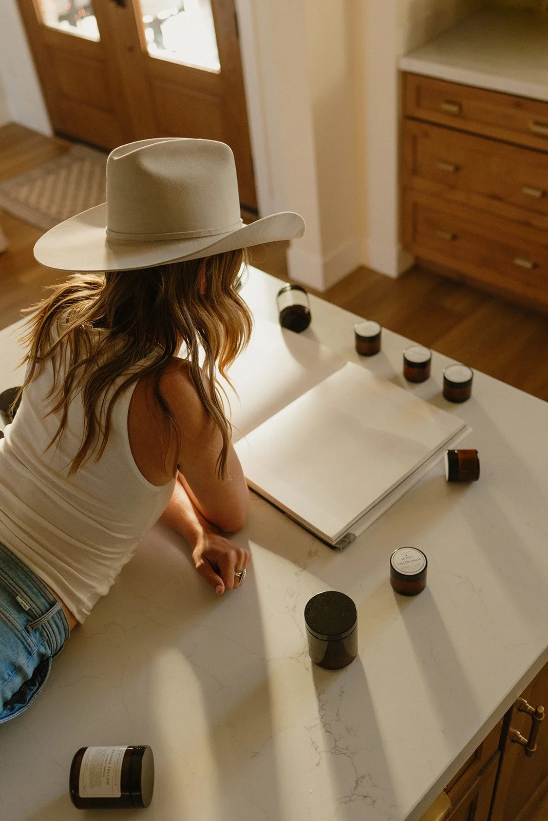 A woman with long hair wearing a white tank top and a beige cowboy hat is leaning on a kitchen countertop, looking at an open notebook surrounded by small amber jars.