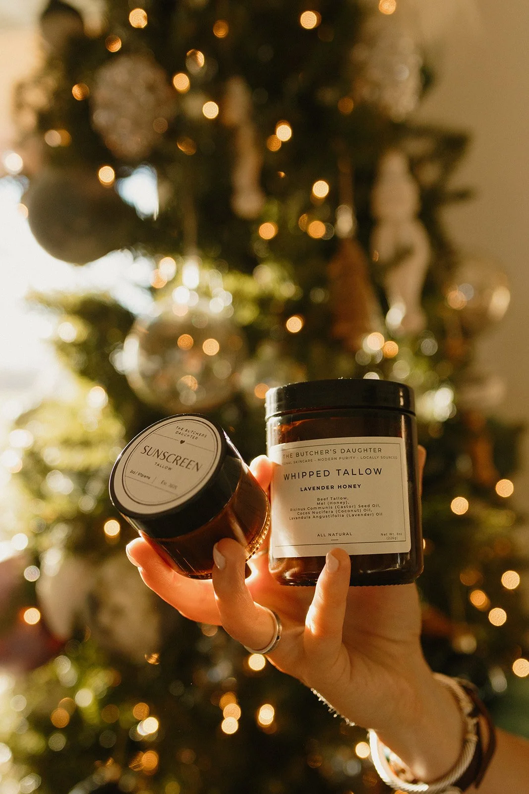 A person holding two natural skincare products called 'Whipped Tallow Lavender Honey' and 'Sunscreen' in front of a decorated Christmas tree with lights.