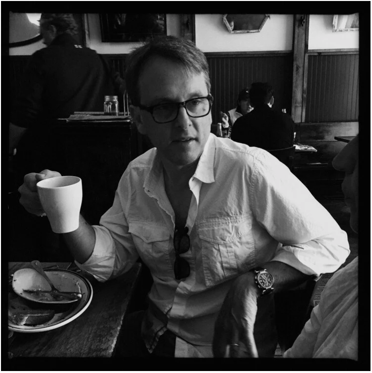 A man with glasses and a wristwatch, wearing a white shirt, sitting at a table in a restaurant, holding a mug of coffee, with a partially eaten plate of food in front of him, engaged in conversation.