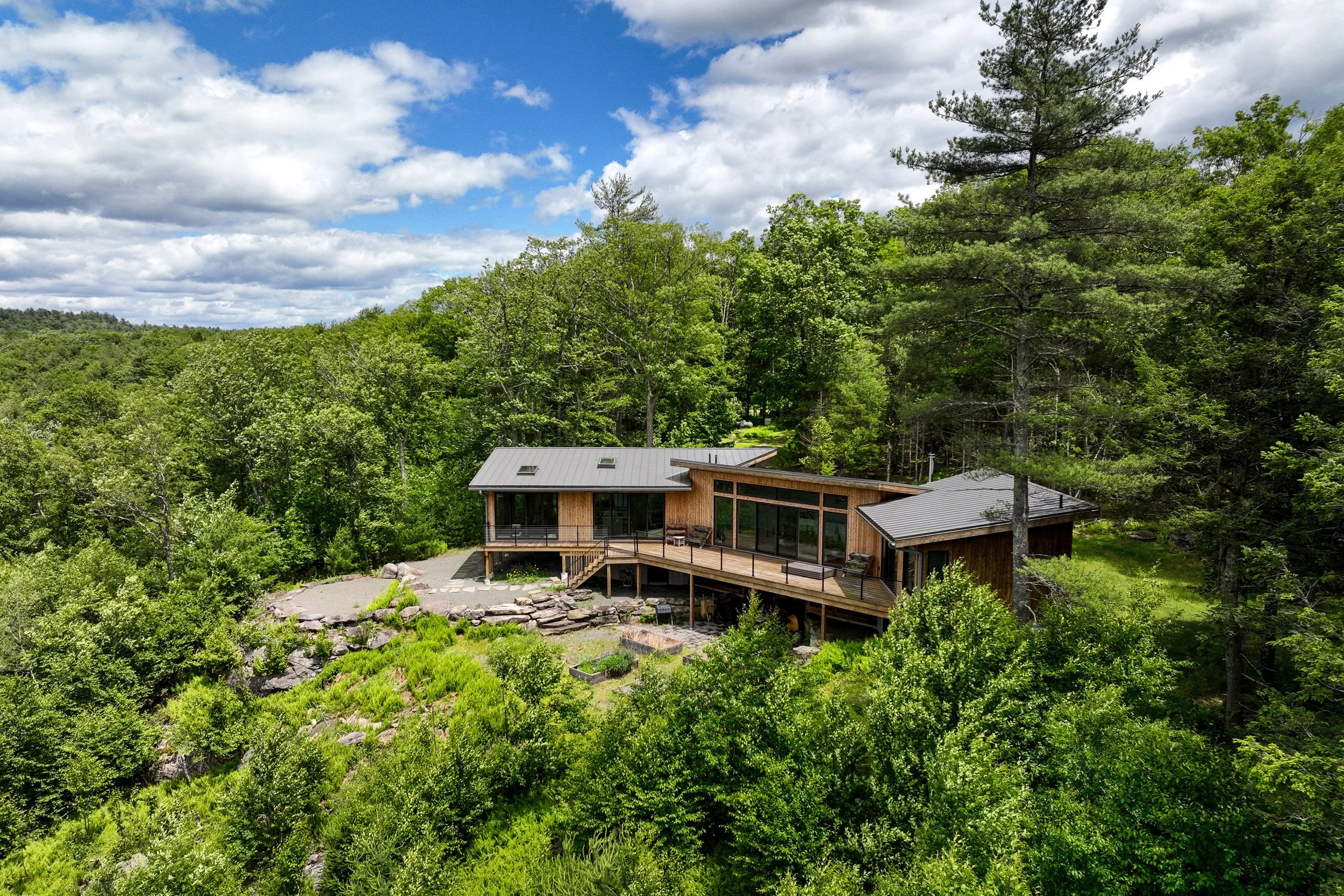 A modern, wooden house with large glass windows, built on a hillside among lush green trees, with a deck and outdoor seating area.
