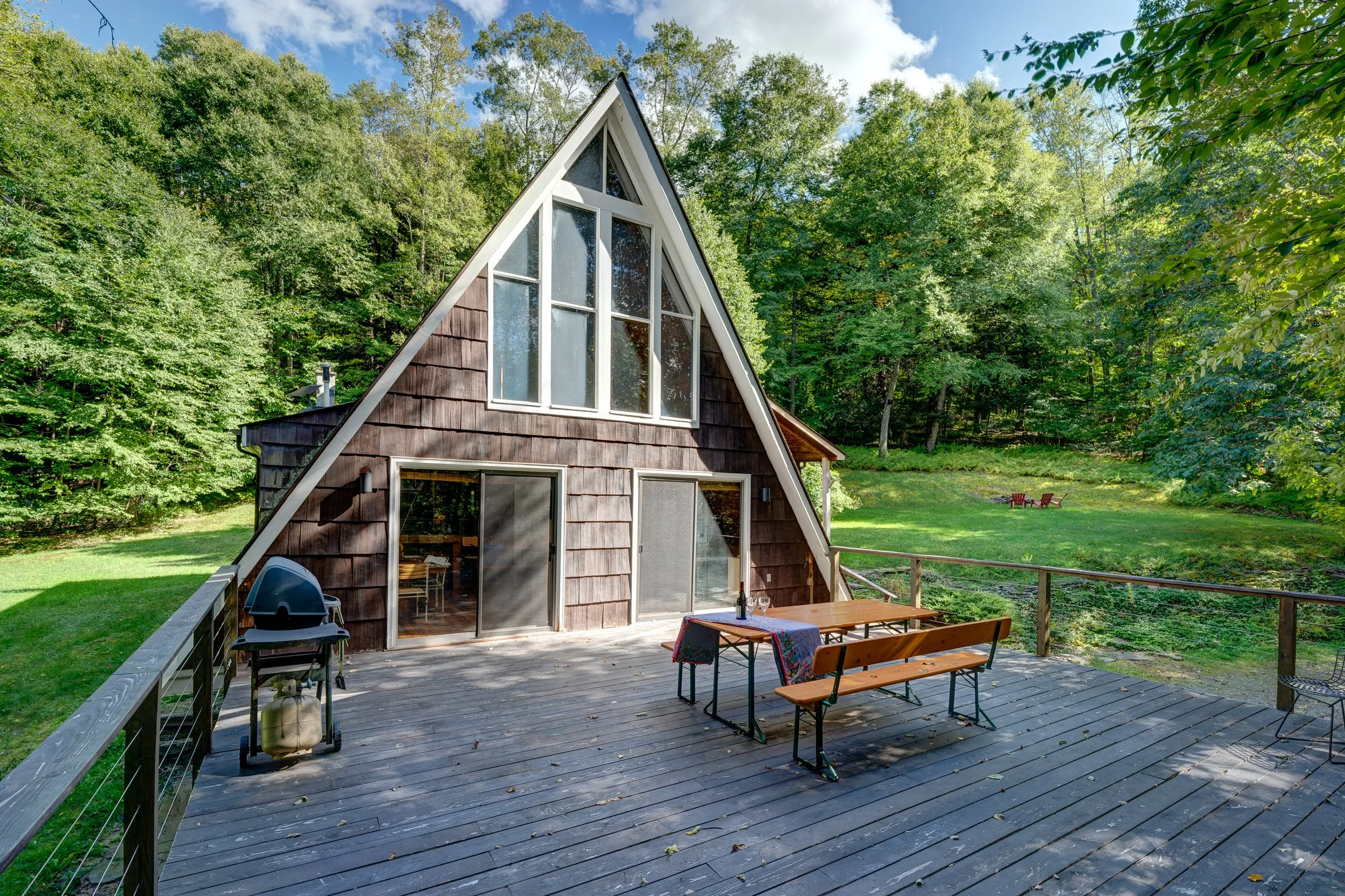 A wooden house with an A-frame architecture, large windows, and a spacious outdoor deck surrounded by trees and green grass.