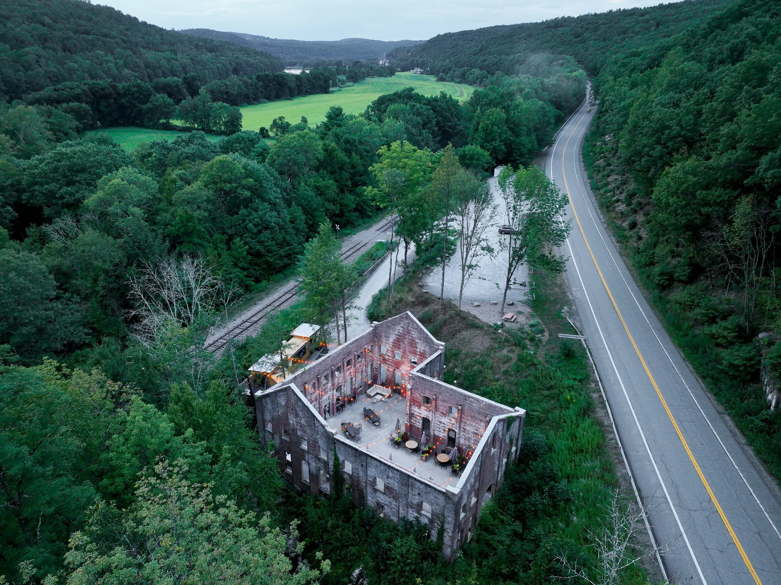 An aerial view of a historic brick building with outdoor seating, surrounded by lush greenery, a railroad track, and a winding road in a hilly rural area.