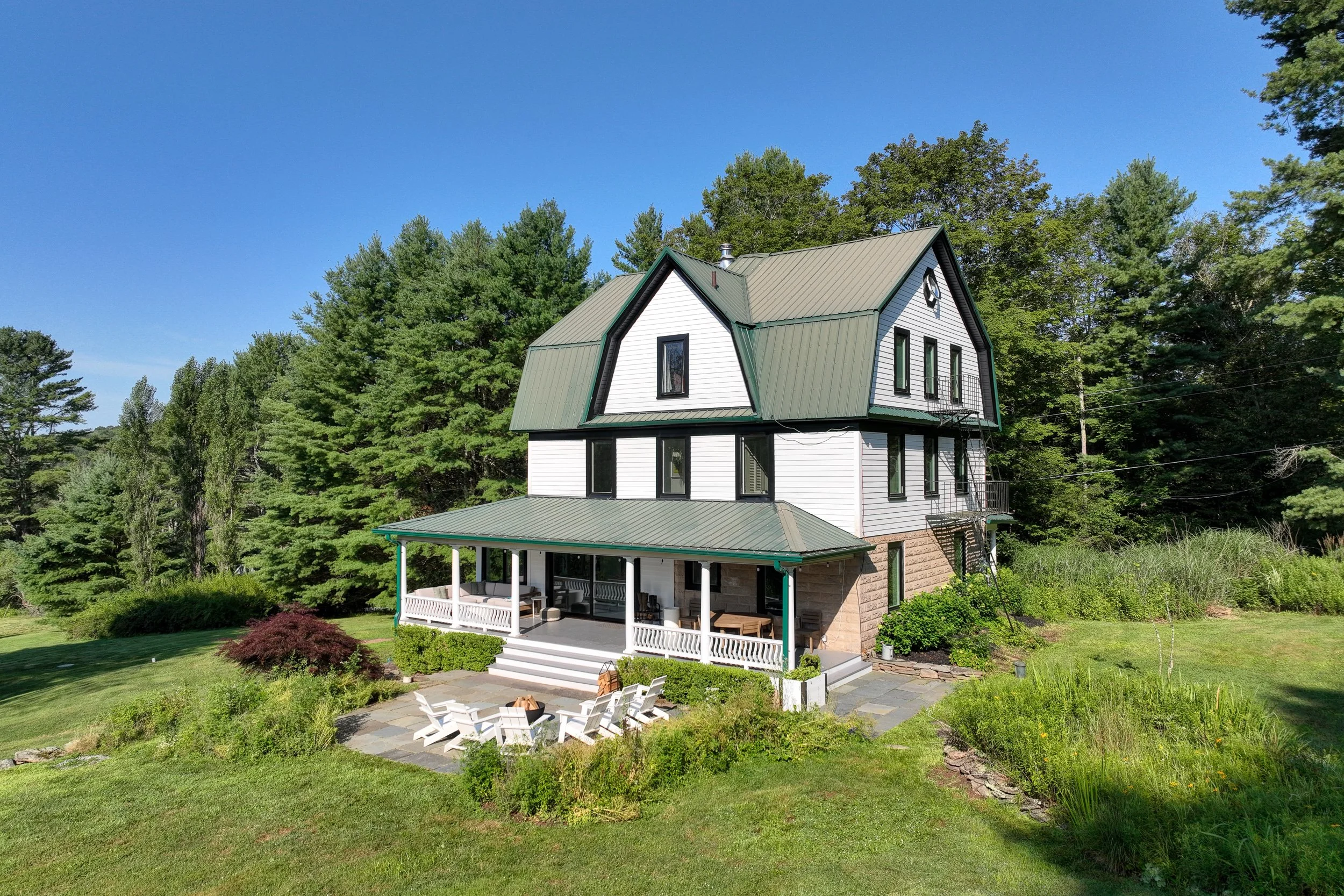 A large white house with a green metal roof and multiple gables, surrounded by green trees and a well-maintained lawn, with a porch and outdoor seating area.