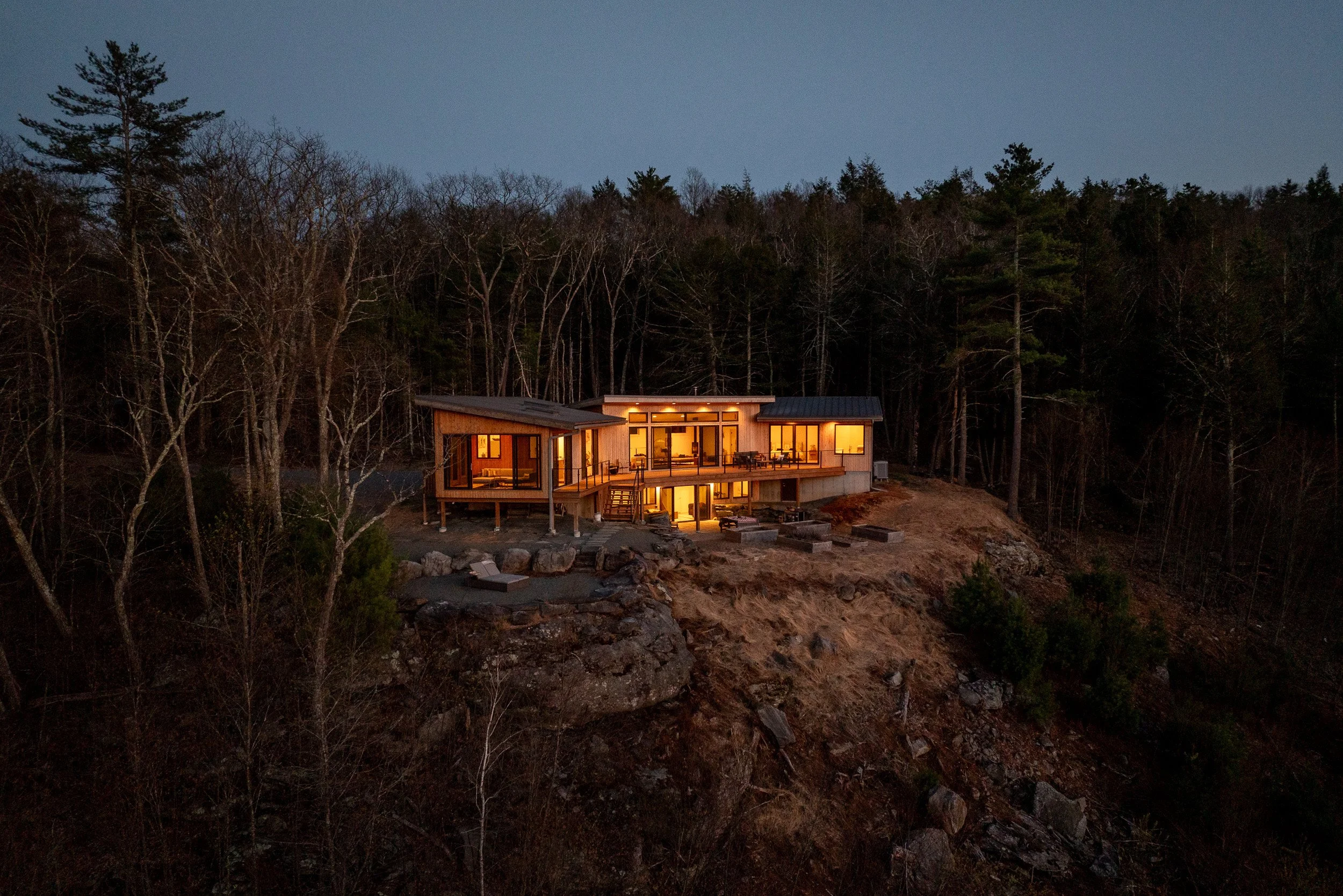 A modern house with large windows at dusk, built on a hillside surrounded by trees, with warm lights glowing from inside.