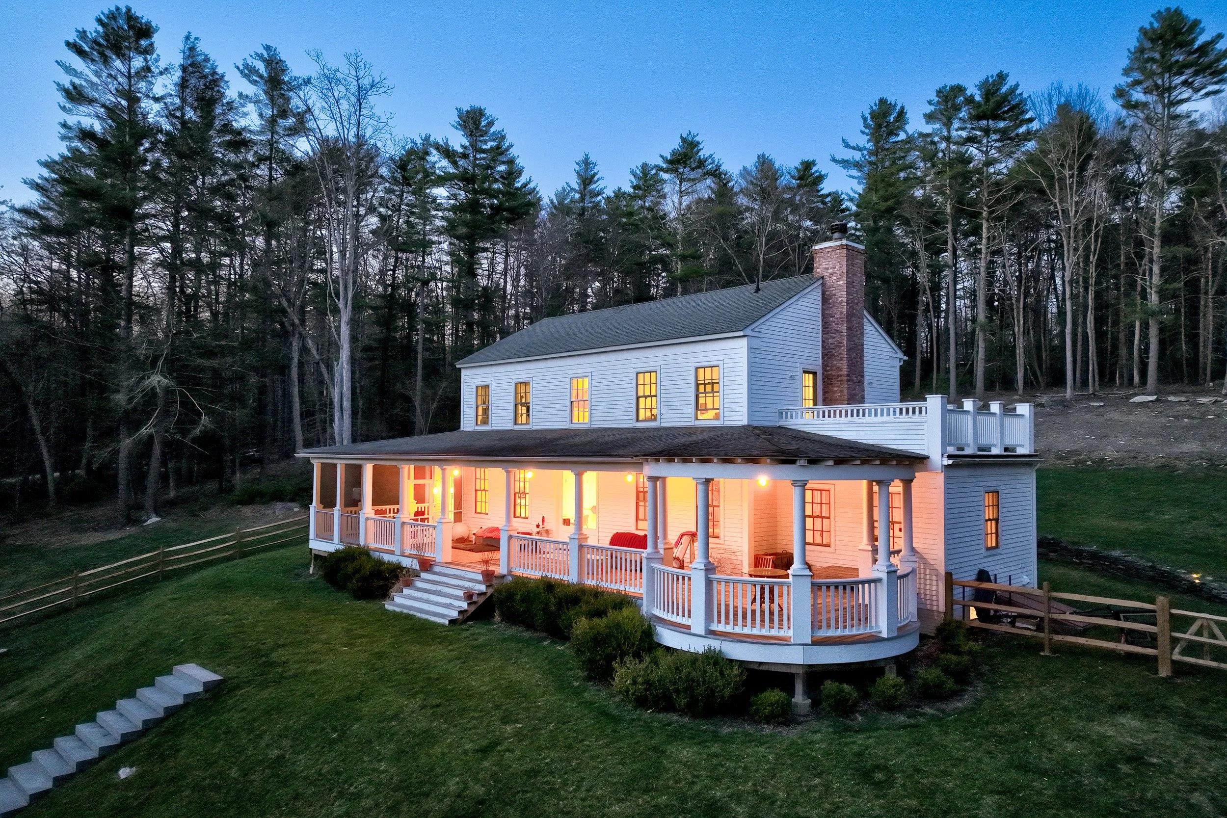 A two-story house with white siding and a chimney, illuminated from inside, surrounded by trees at dusk.