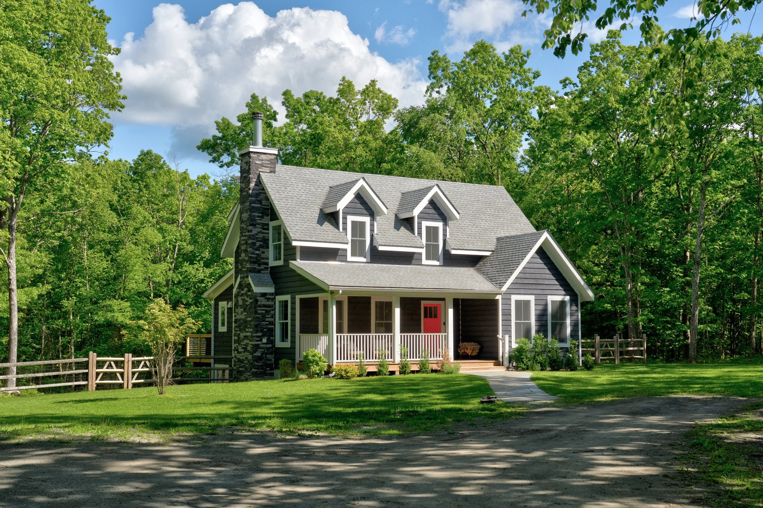 A house with dark siding, white trim, and a gray roof, surrounded by green trees and a lawn, with a dirt driveway in the front.