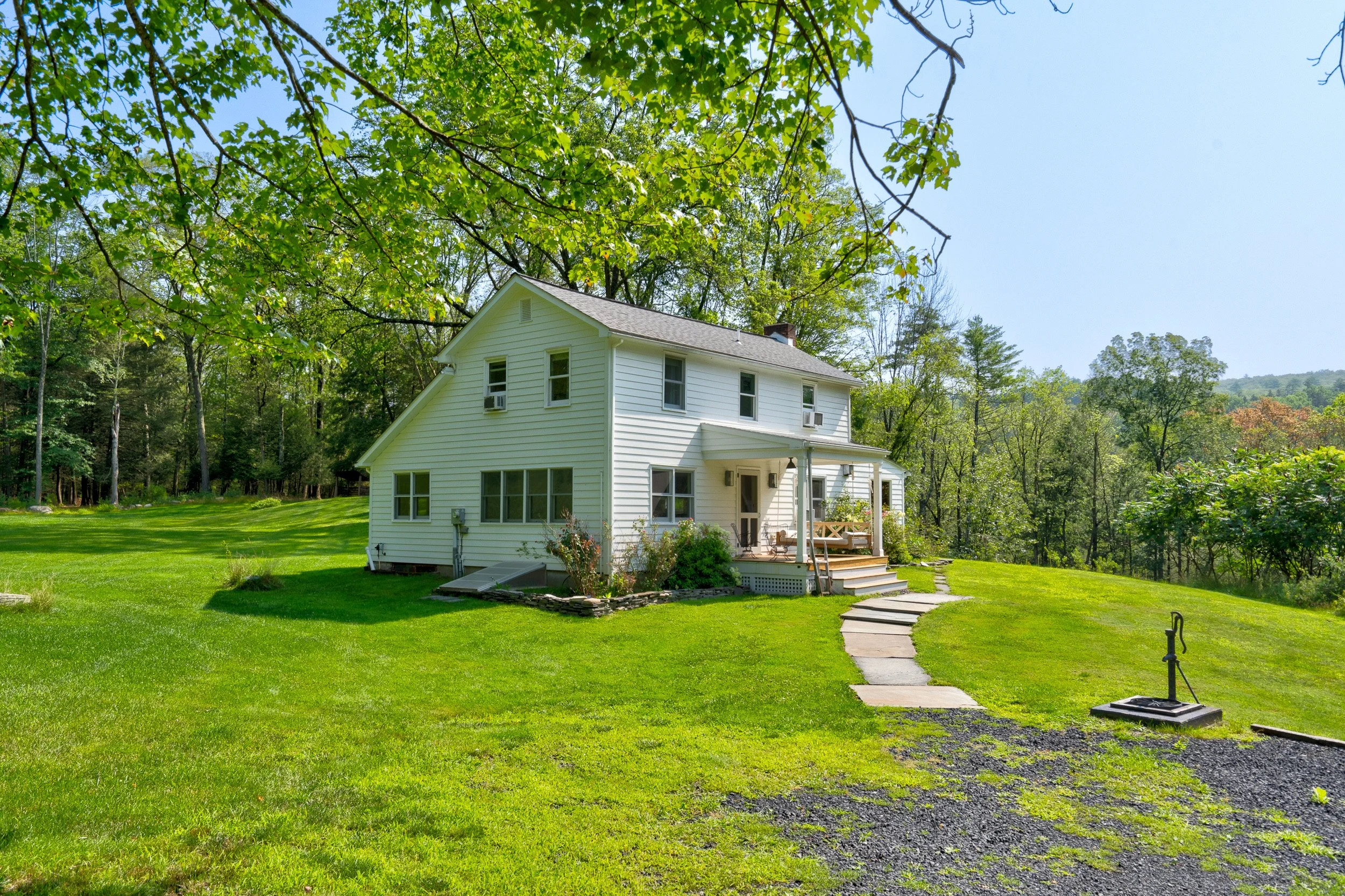 A white two-story house with a porch, surrounded by green grass and trees, with a stone pathway leading to the porch, under a bright blue sky.