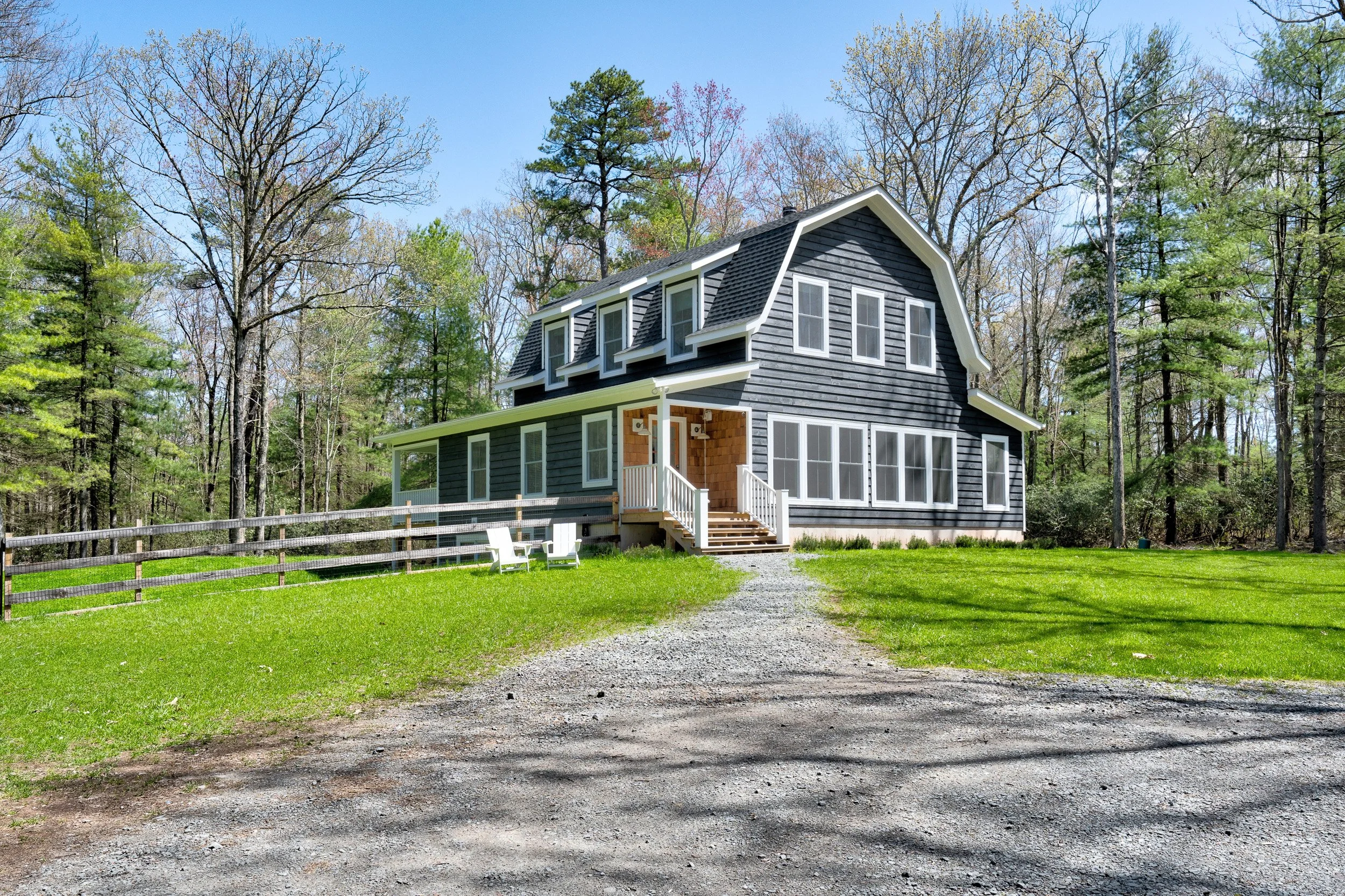 A black two-story house with white trim, surrounded by trees, with a gravel driveway leading to the front porch with stairs and two white chairs.