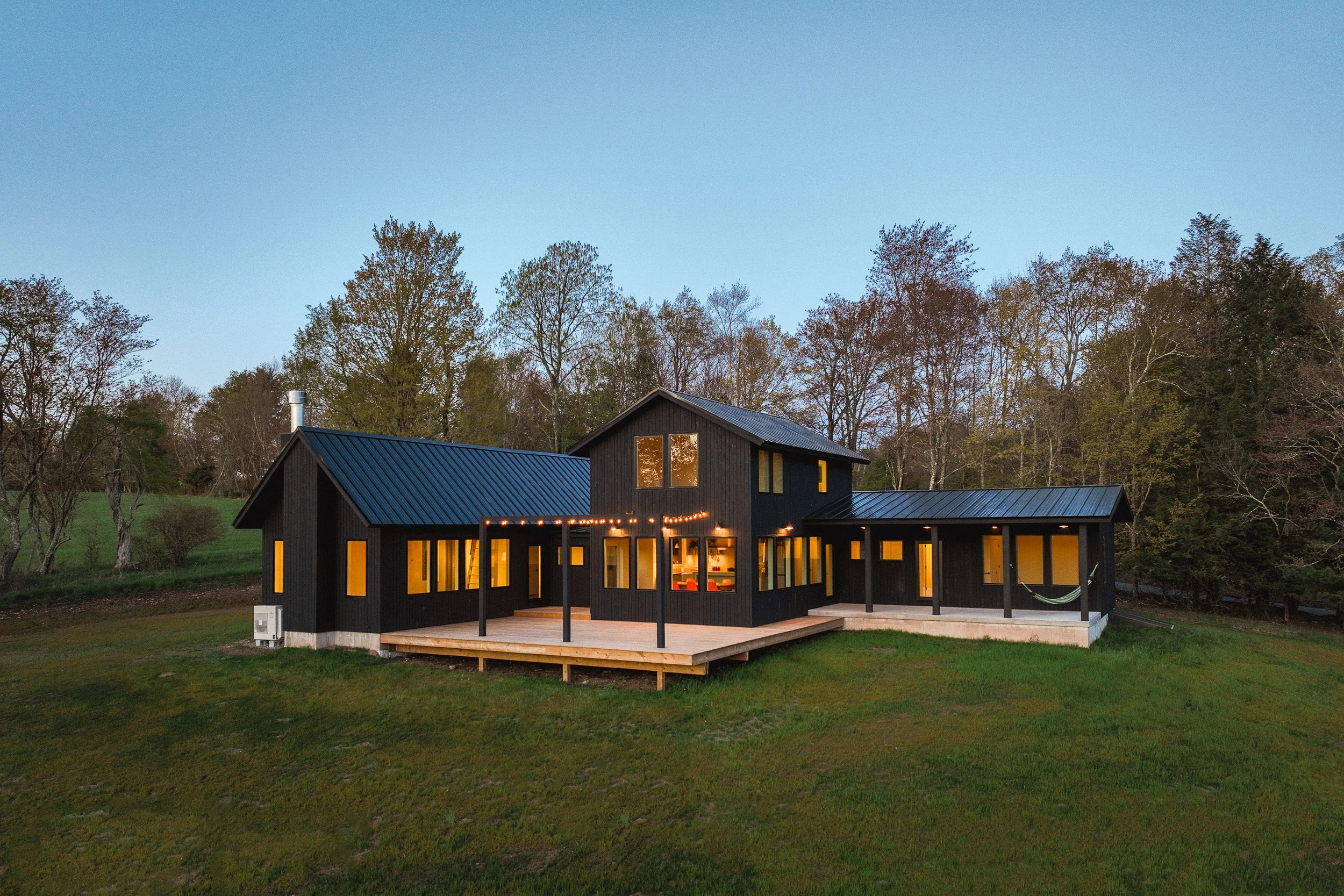 A newly built modern, contemporary house with wood siding painted black. The porch has string lights and wraps around to a dining space with a hammock. 