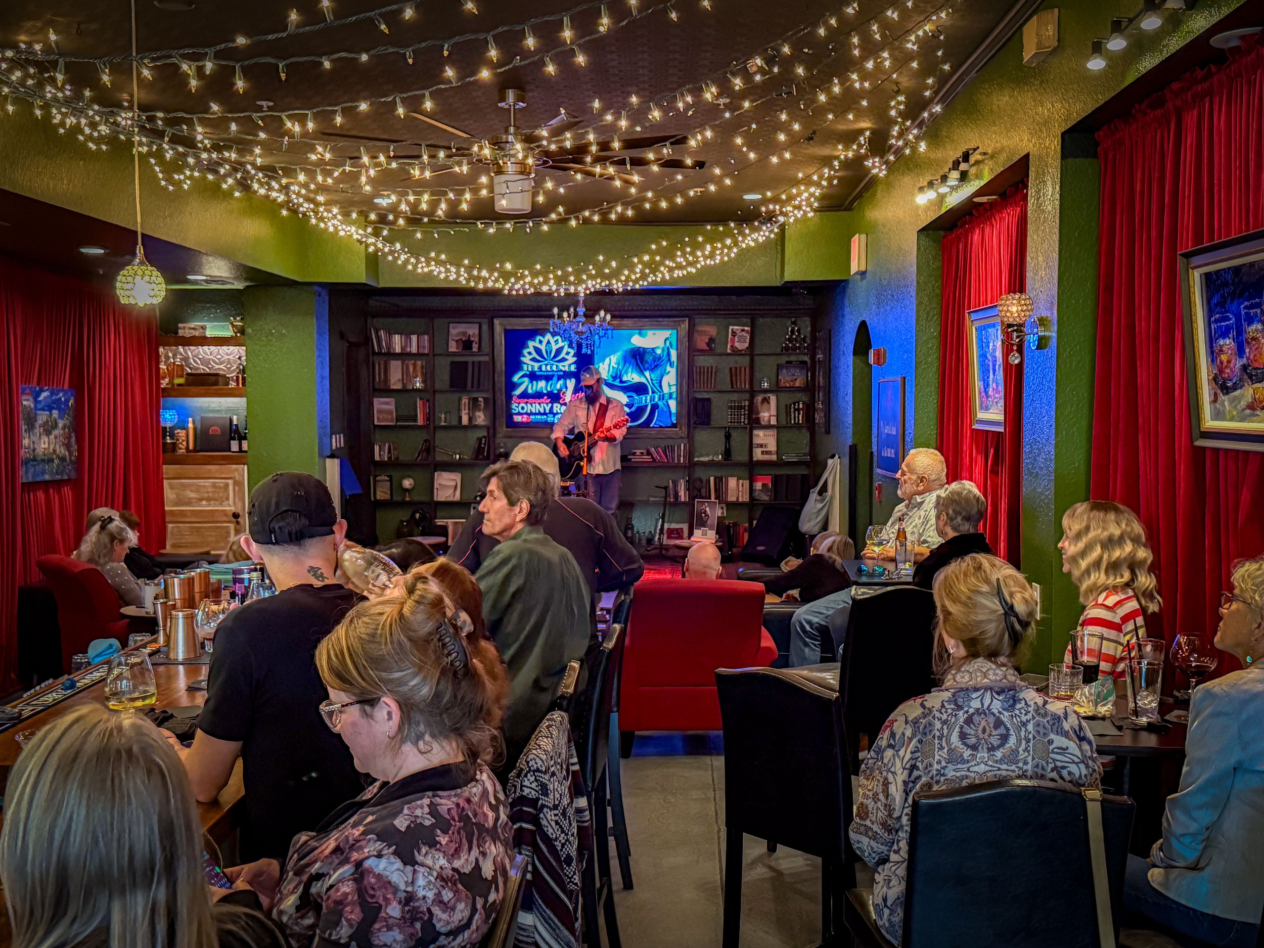 A live music performance at an intimate venue with customers seated at tables. The stage has a performer playing guitar, with a neon sign and bookshelf in the background. The room is decorated with string lights, red curtains, and framed artwork on g