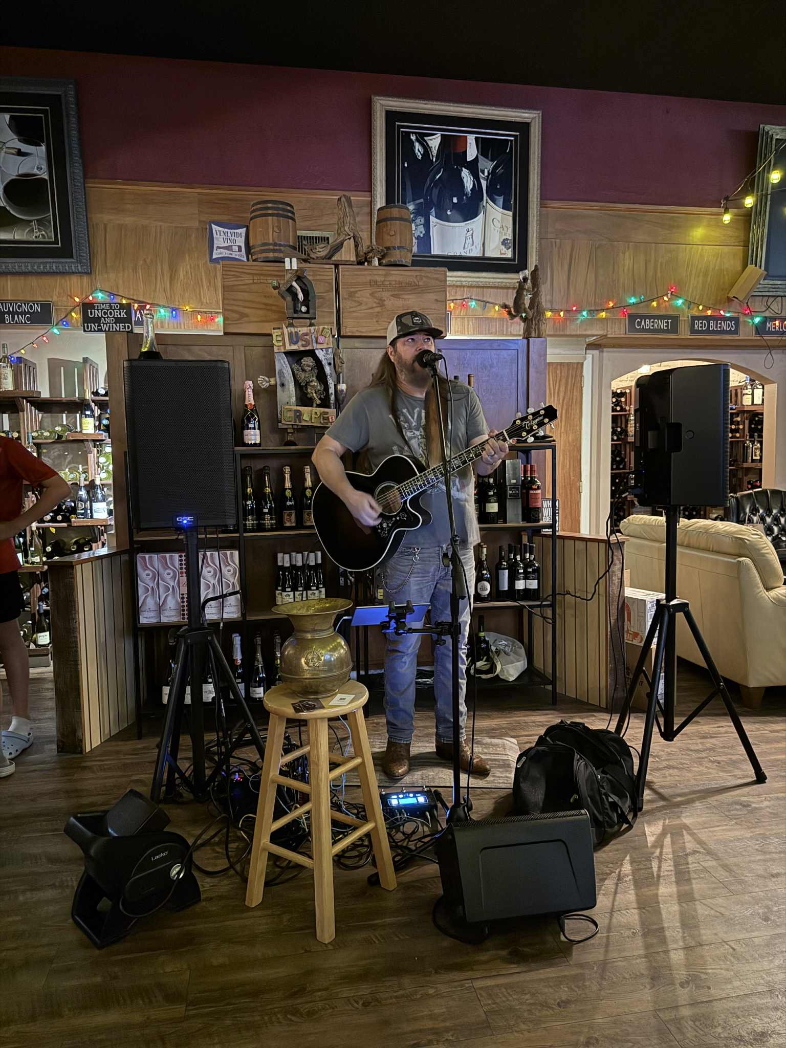 A musician performs with an acoustic guitar on a stage set up inside a restaurant or bar, with beer bottles on shelves behind him and colorful string lights overhead.