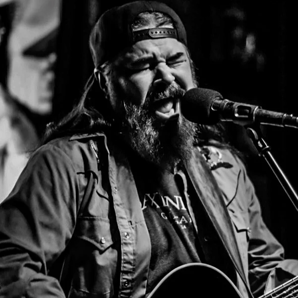 A man with a beard and long hair wearing a backwards cap singing into a microphone while playing an acoustic guitar.