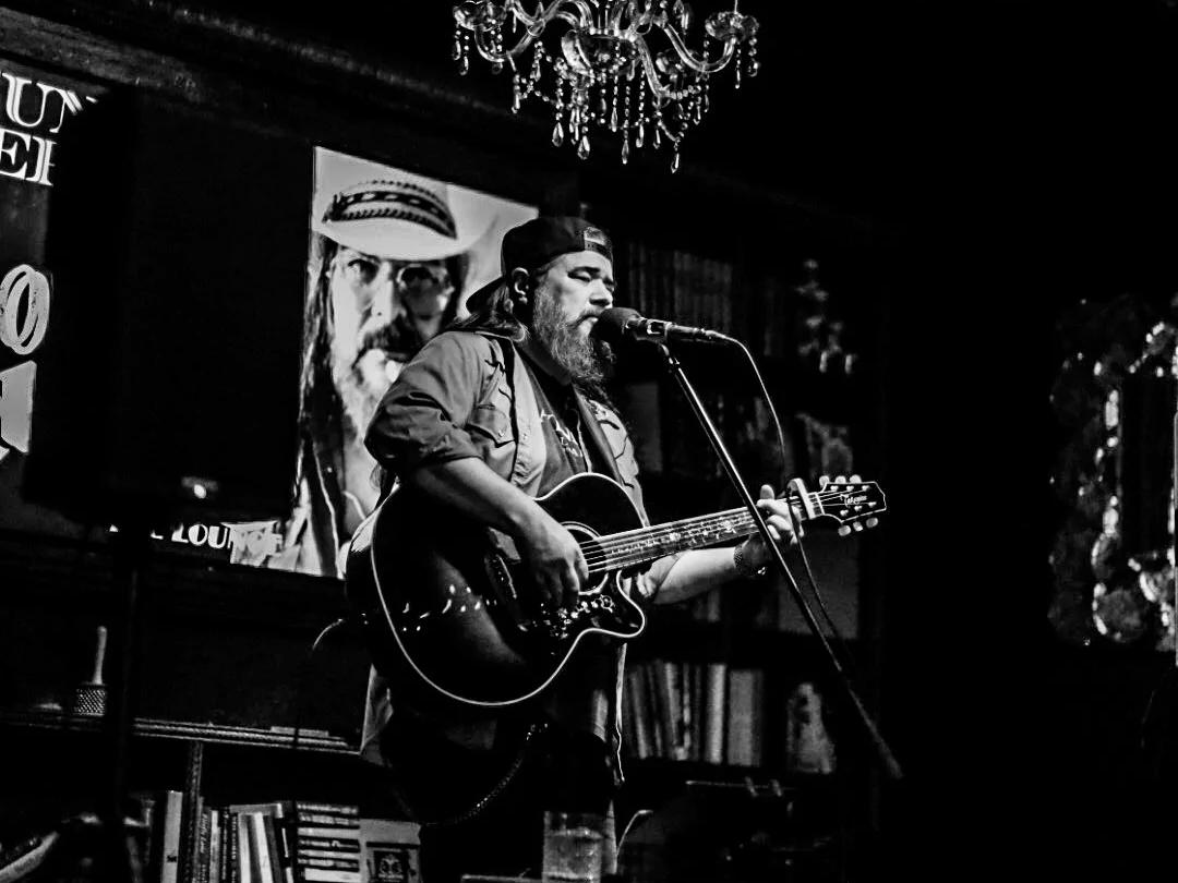 A male musician with long hair and a beard wearing a cap playing an acoustic guitar and singing into a microphone on stage, with a large screen behind him displaying a portrait of himself, and a chandelier hanging from the ceiling.