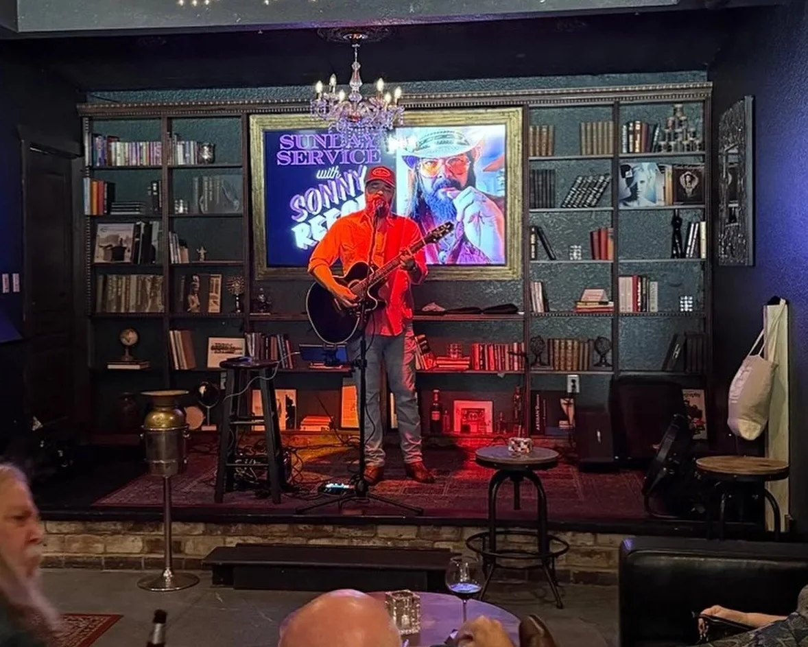 Man performing on stage with guitar in a cozy bar or cafe, with shelves of books and framed photos behind him, and a large screen displaying a promotional graphic for the event. Audience members seated in front.