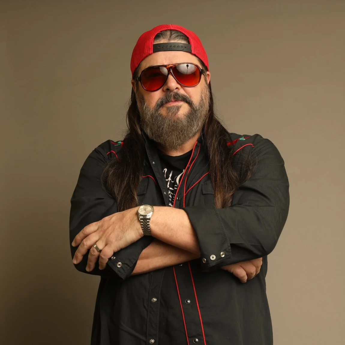 A man with long hair and a beard wearing a red baseball cap backwards, sunglasses, a black shirt with red accents, a watch, and rings, standing with arms crossed against a plain background.