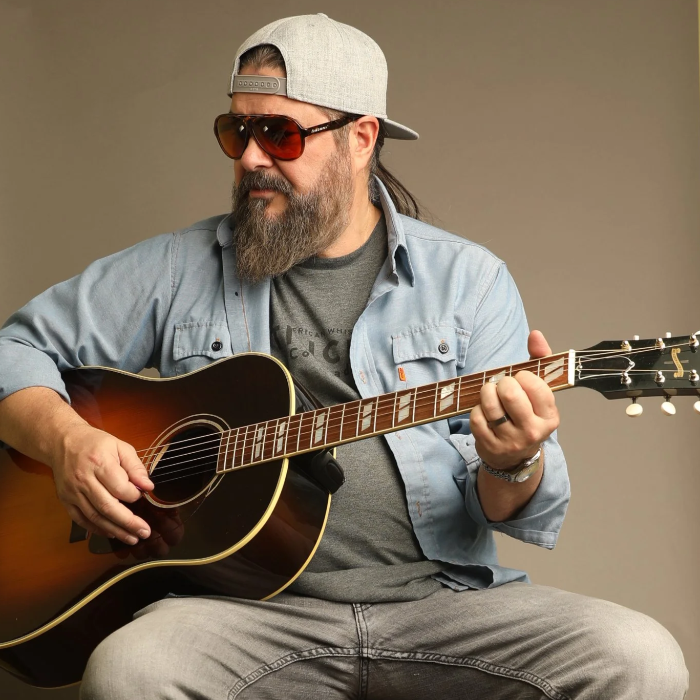 A man with a beard, sunglasses, a gray baseball cap worn backwards, a gray t-shirt, and a light blue button-up shirt playing an acoustic guitar.