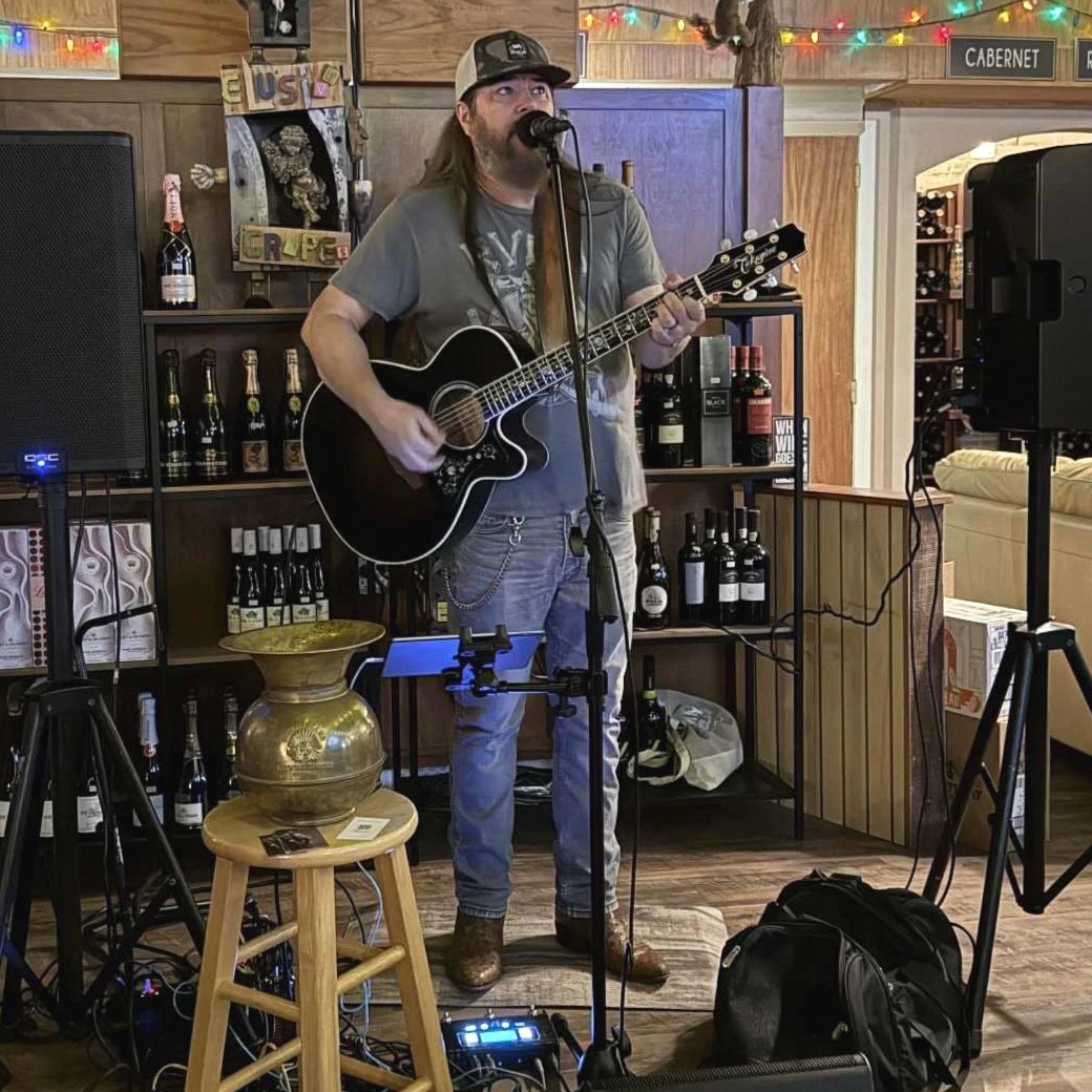 A musician performs with an acoustic guitar on a stage set up inside a restaurant or bar, with beer bottles on shelves behind him and colorful string lights overhead.