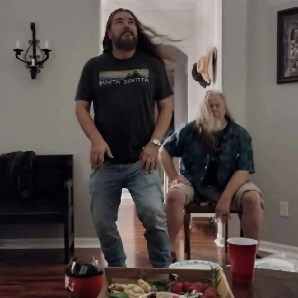 Sonny Reece & Kim Groff from Keltstache Music at a social gathering, one standing with long hair and beard, wearing a South Dakota t-shirt, and the other sitting on a chair with long blond hair, wearing a denim shirt. There are snacks, drinks, and a 