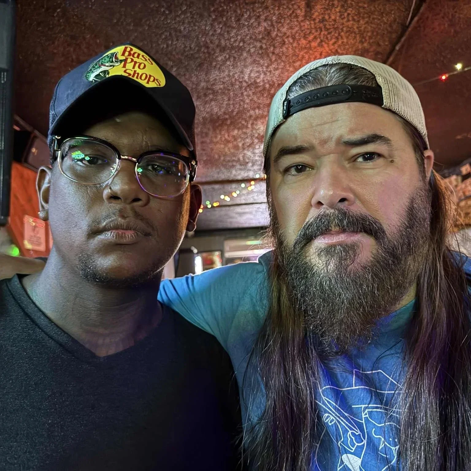 Two men standing close together inside a dimly lit bar or restaurant, with string lights hanging from the ceiling. One man is wearing glasses, a black cap with a baseball logo, and a black shirt. The other man has long hair, a beard, and is wearing a