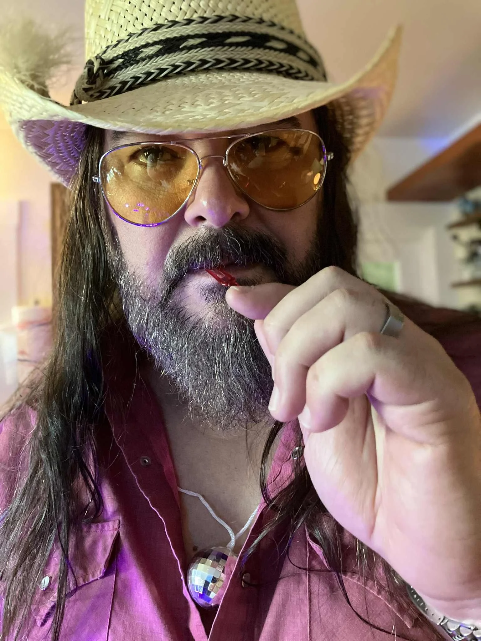 A man wearing a straw hat with black and white bands, yellow-tinted aviator sunglasses, a pink shirt, and a silver disco ball necklace, holding a lollipop near his lips in an indoor setting.