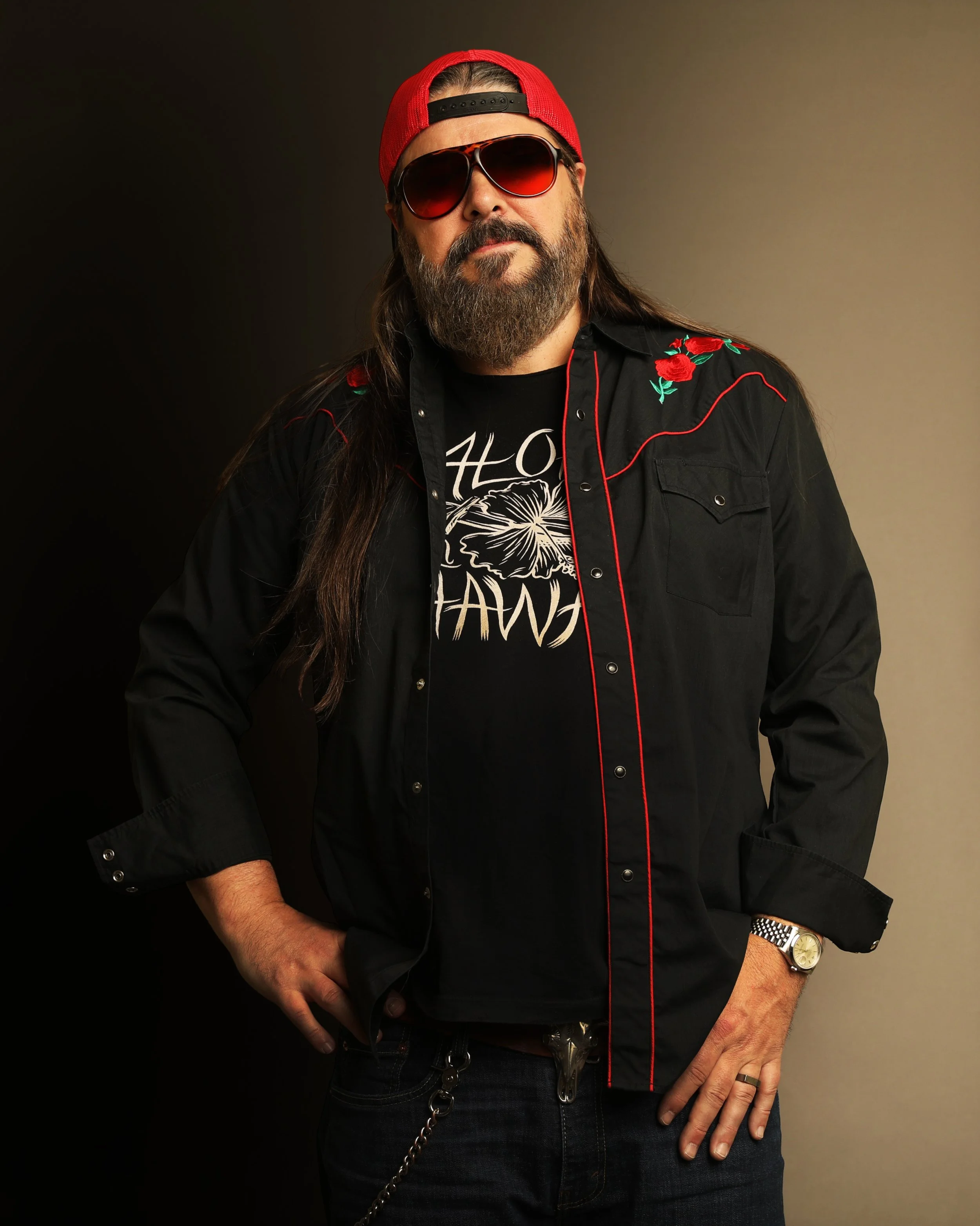 Sonny Reece with long hair and a beard wearing sunglasses, a red baseball cap backwards, a black pearlsnap shirt with a graphic and the words 'Aloha Hawaii', and a black jacket with red embroidery. He is standing with one hand on his hip against a da