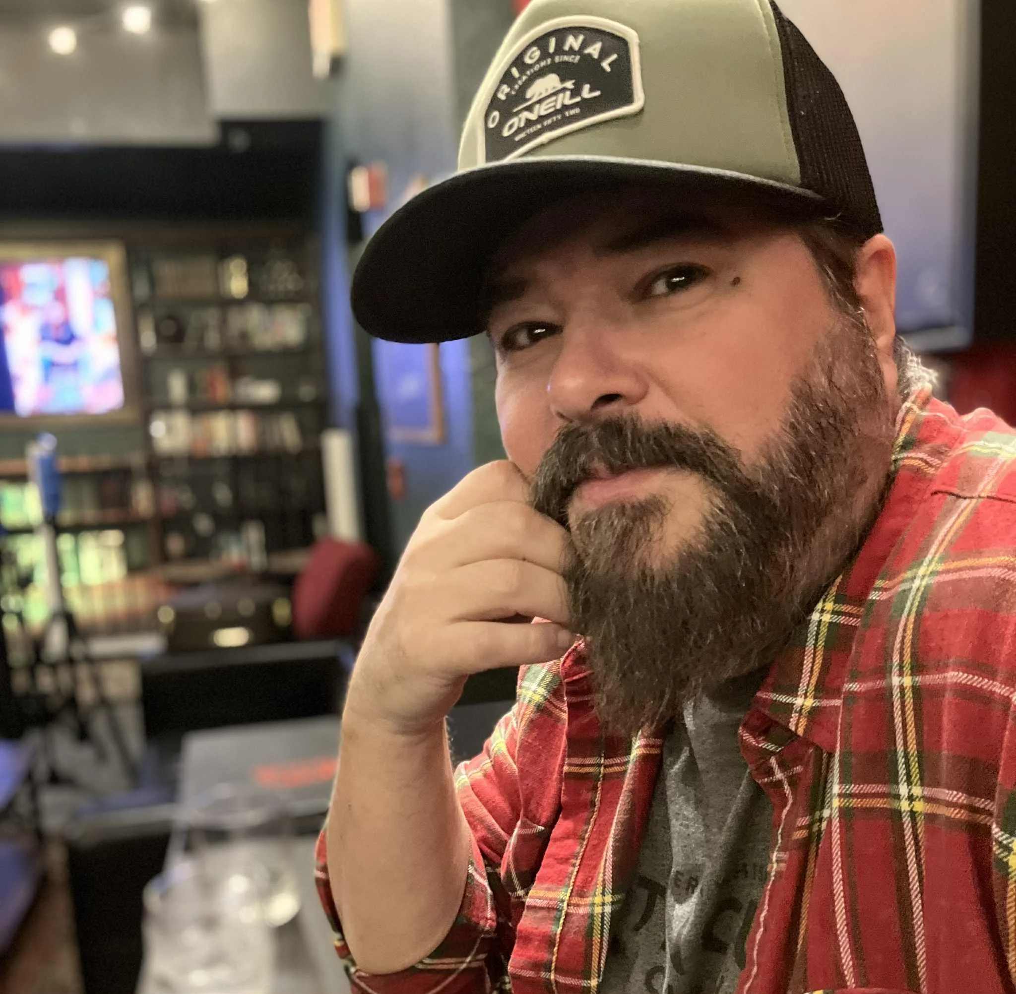 A man with a beard and mustache wearing a baseball cap and a red plaid shirt, sitting at a bar or restaurant.