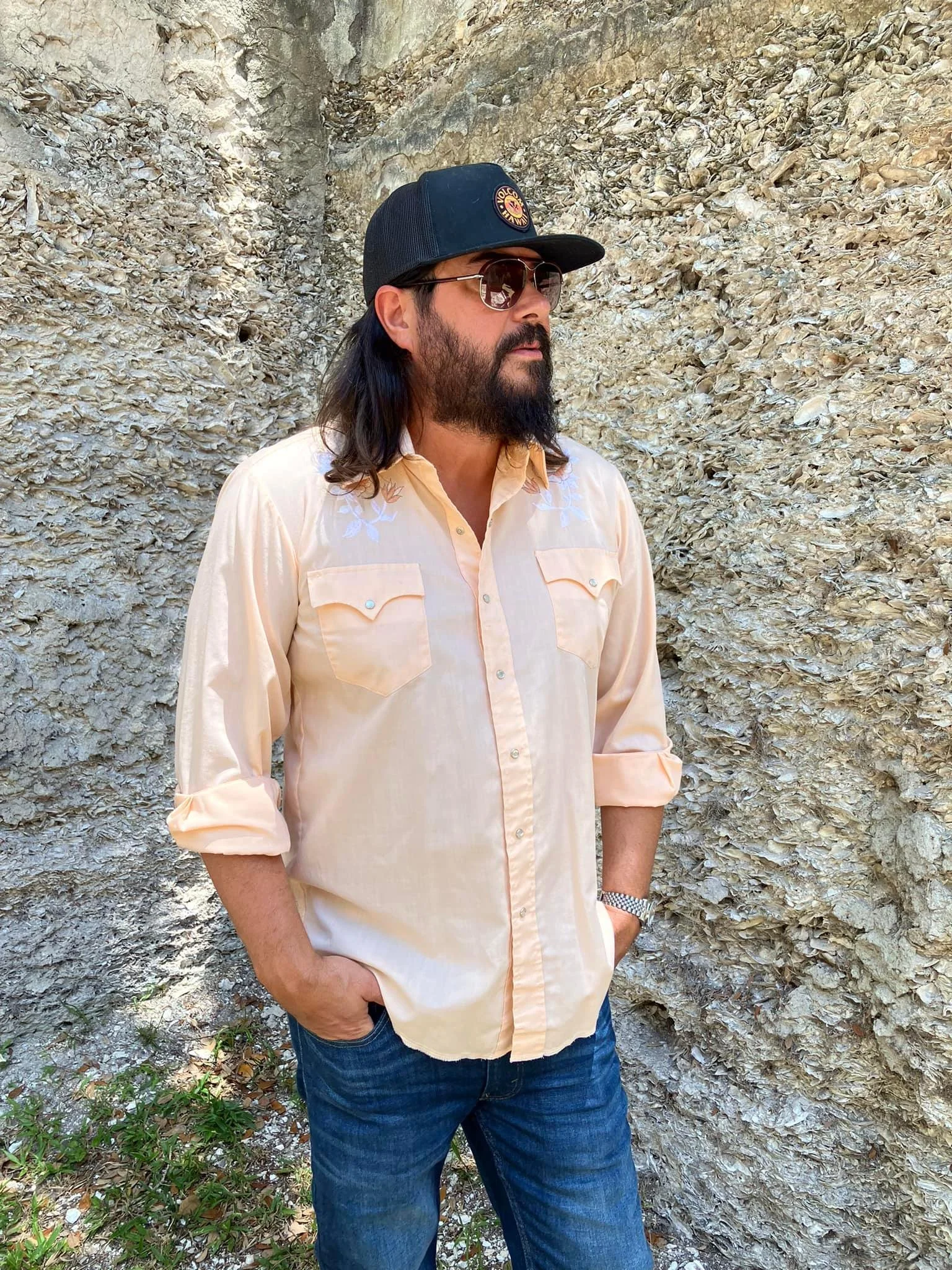 A man with long dark hair and beard wearing sunglasses, a black cap, a cream-colored button-up shirt with rolled-up sleeves, and jeans, standing outdoors against a rocky background with his hands in his pockets.