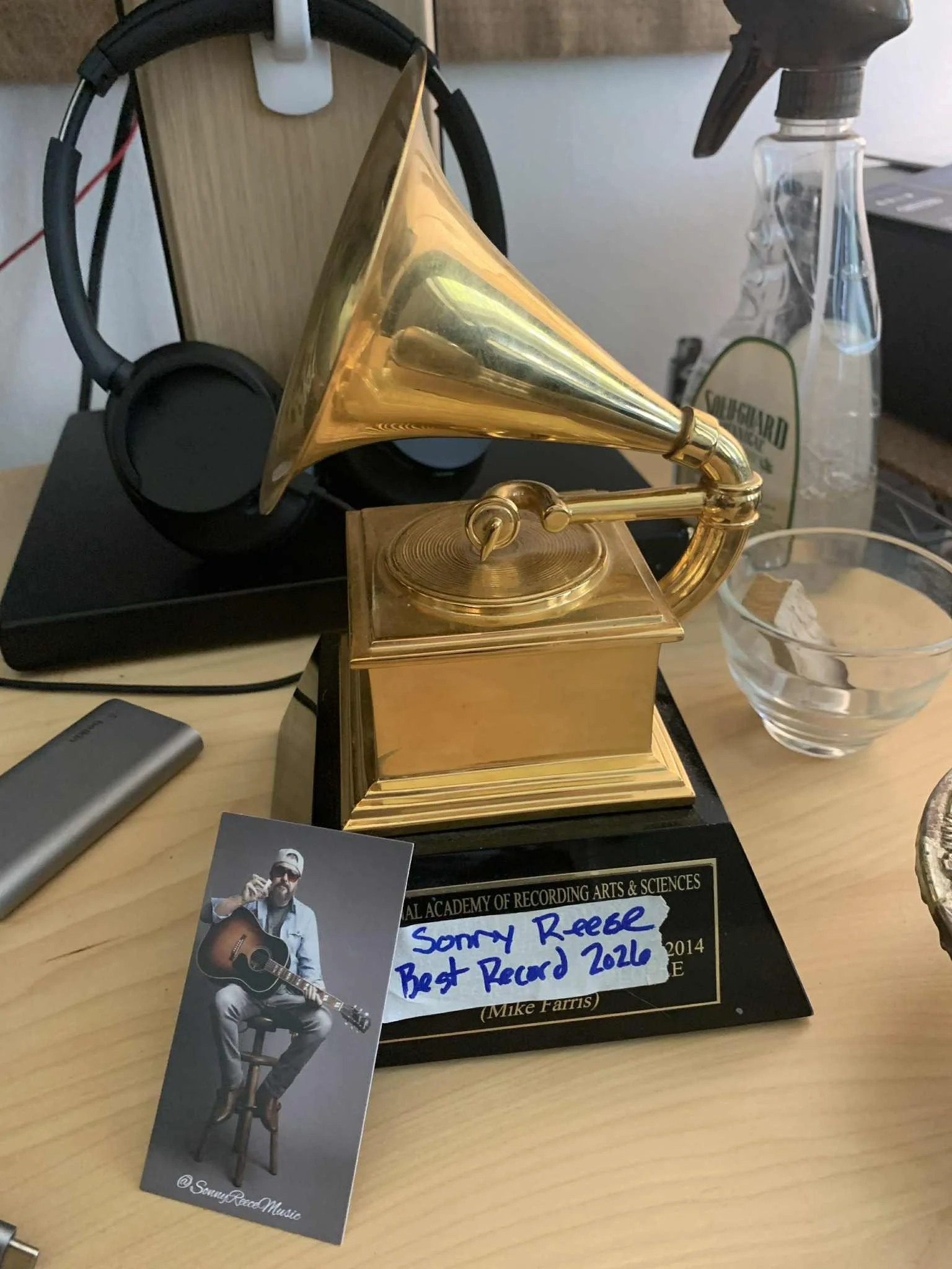 Golden gramophone trophy on a desk with a handwritten note saying 'Sonny Reece Best Record 2020', a pair of black headphones, a business card of a man with a guitar, and various office items.
