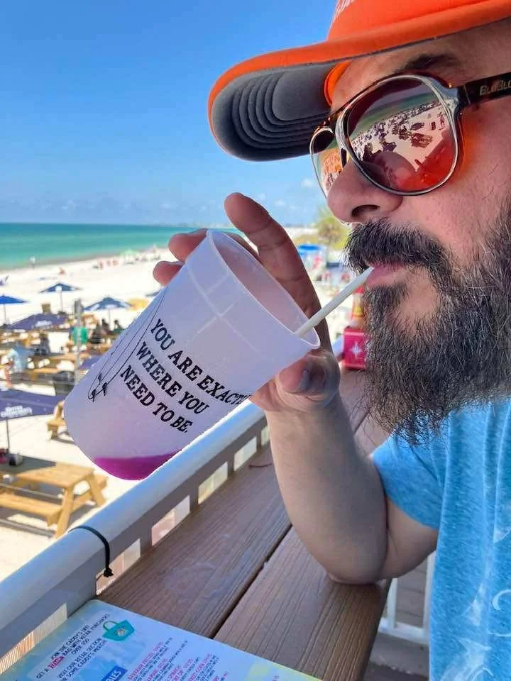 A man wearing a large orange sun hat and sunglasses with reflective lenses is drinking from a large white cup with a straw, on a beachside balcony with beach chairs and umbrellas, ocean waves, and a clear blue sky in the background.
