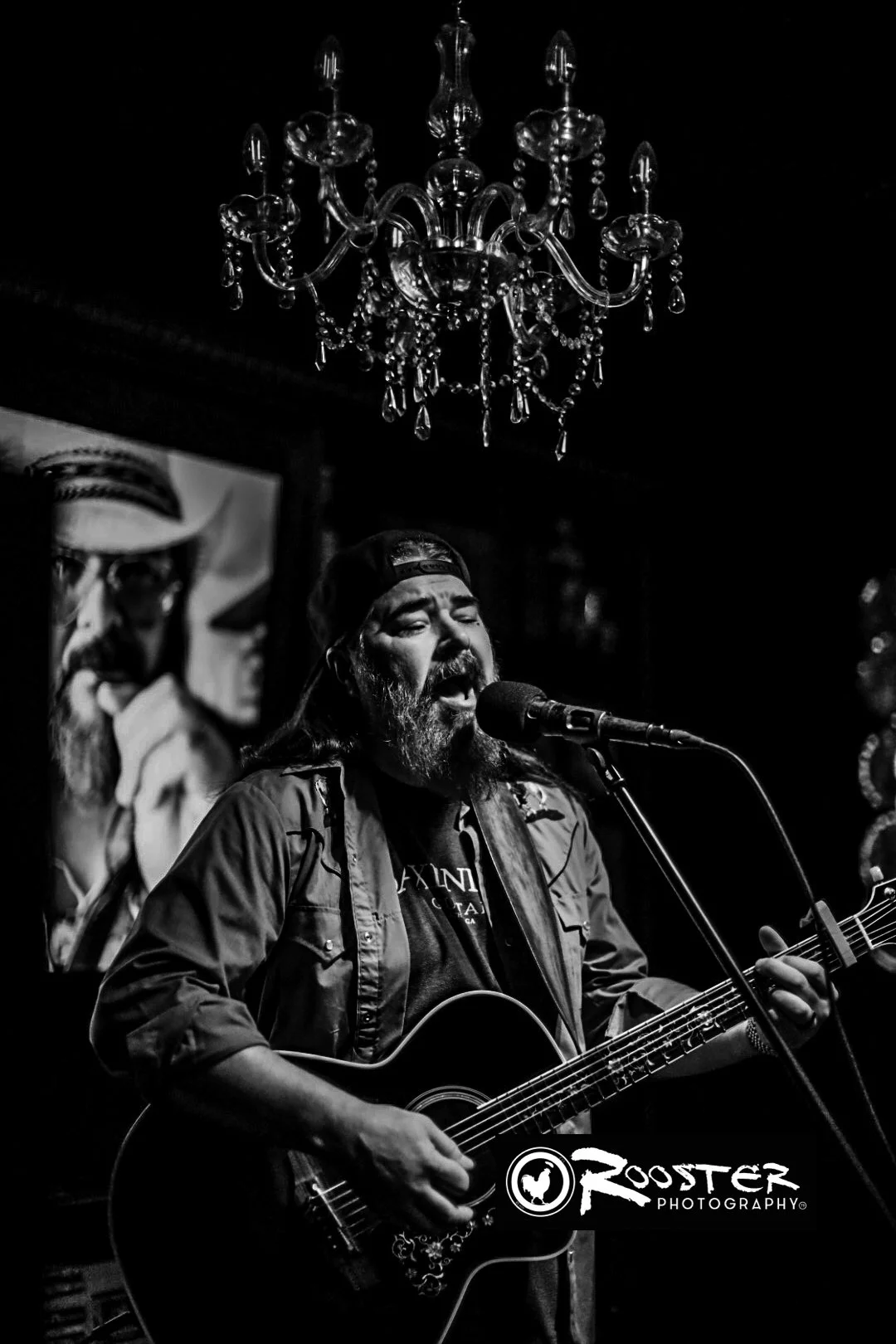 Black and white photo of a man singing into a microphone while playing an acoustic guitar, with a chandelier hanging above him and a poster in the background.