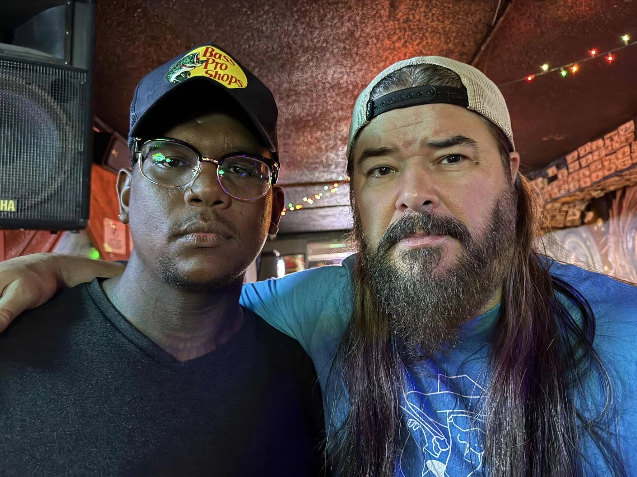 Two men standing close together inside a dimly lit bar or restaurant, with string lights hanging from the ceiling. One man is wearing glasses, a black cap with a baseball logo, and a black shirt. The other man has long hair, a beard, and is wearing a