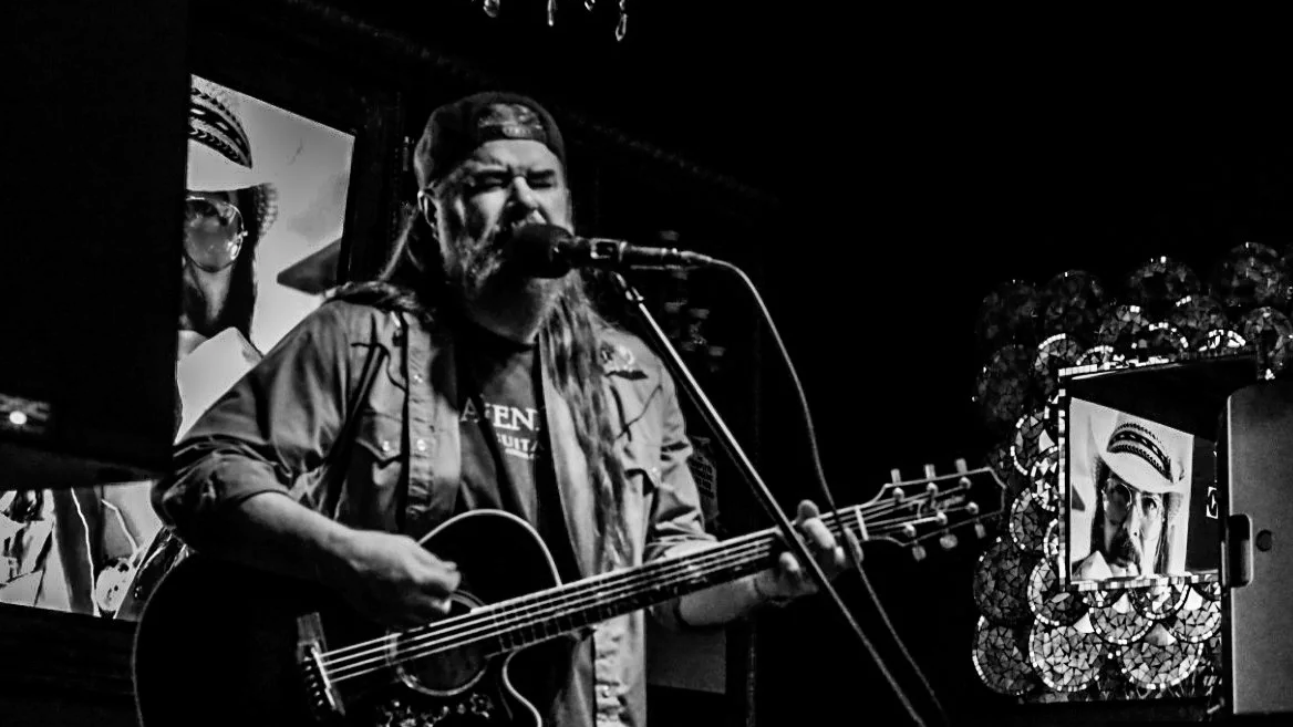 A man with long hair, a beard, wearing a cap backwards, is singing into a microphone while playing an acoustic guitar. There are framed pictures and decorations in the background.
