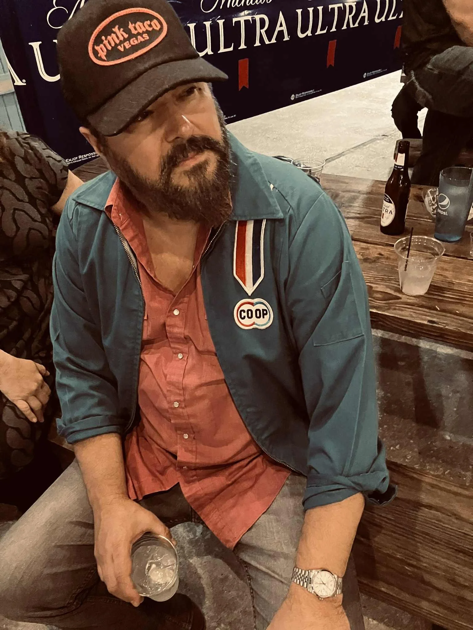 A man with a beard and mustache wearing a cap with 'Pink Taco Vegas' embroidered on it, a blue jacket with a 'COOP' patch, a coral button-up shirt, and a silver watch, sitting at a wooden table with drinks around him in a bar or restaurant setting.