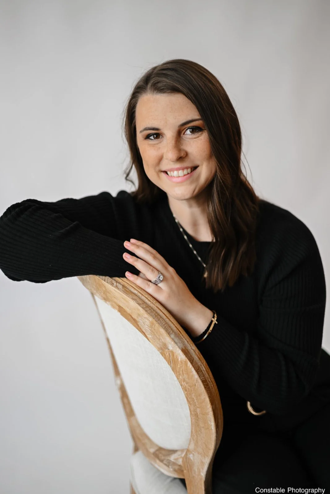 A woman with long brown hair wearing a black sweater, smiling, resting her arm on the back of an upholstered chair with a wooden frame, against a plain light gray background.