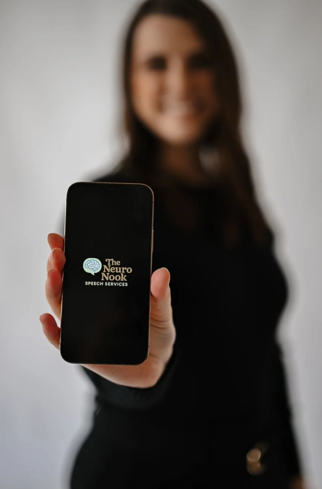 A woman holding a smartphone displaying a logo for 'The Neuro Nook Speech Services' against a plain background.
