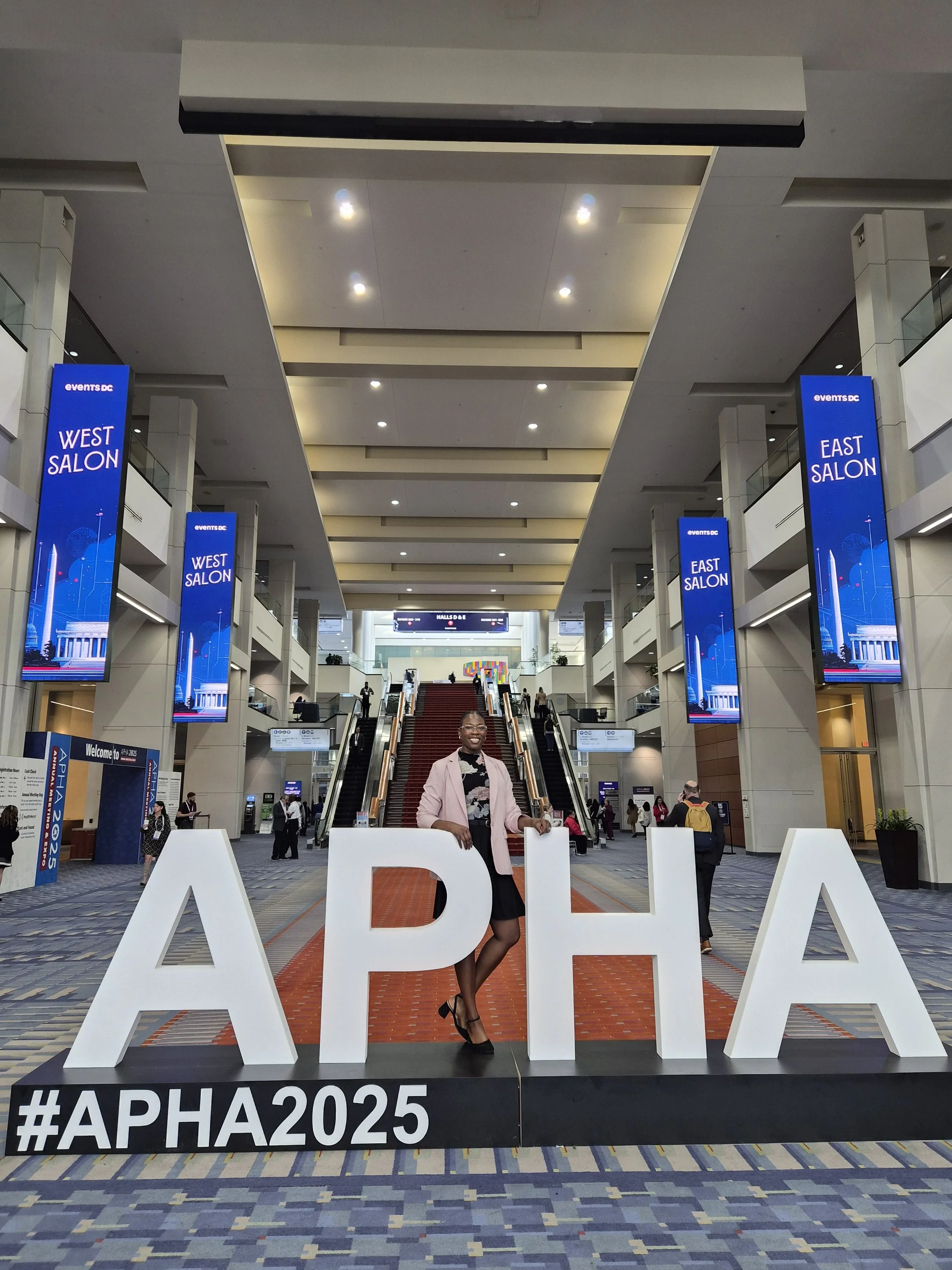 A woman in a pink blazer and black dress posing behind large white letters spelling 'APIA' with a hashtag #APA2025 at an indoor convention center. Digital screens display 'East Salon' and 'West Salon,' and escalators are visible in the background.