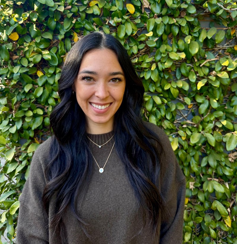 A woman with long dark wavy hair smiling in front of a green leafy hedge, wearing a brown sweater and layered necklaces.