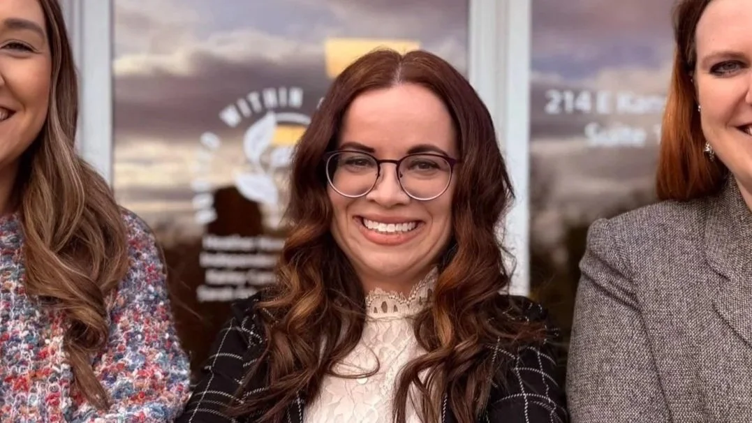 A woman with long dark hair, wearing a black top and a purple beaded necklace, smiling and looking at the camera.