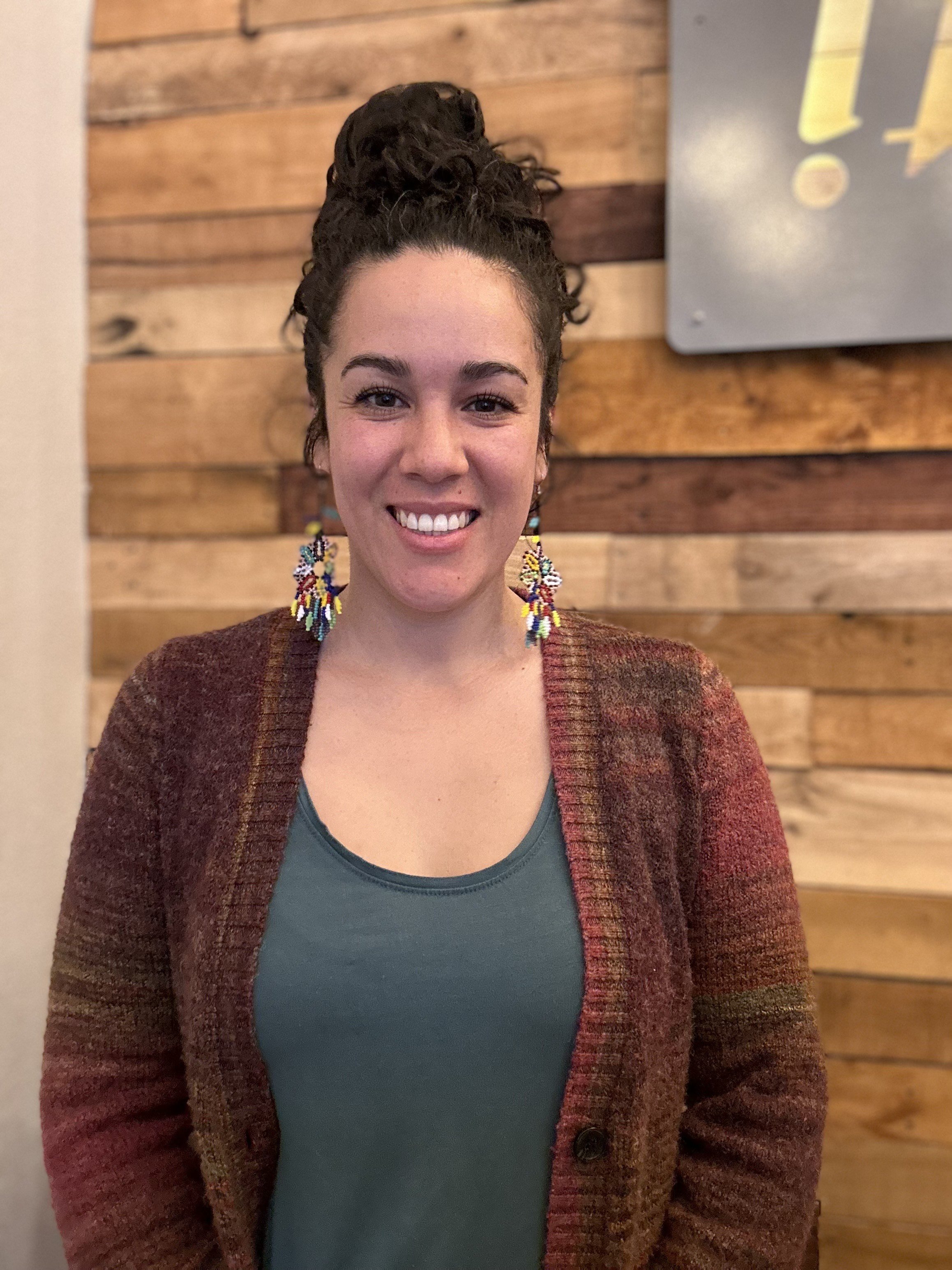 A smiling woman with dark curly hair styled in a bun, wearing colorful beaded earrings and a brown cardigan over a teal top, standing in front of a wooden wall.