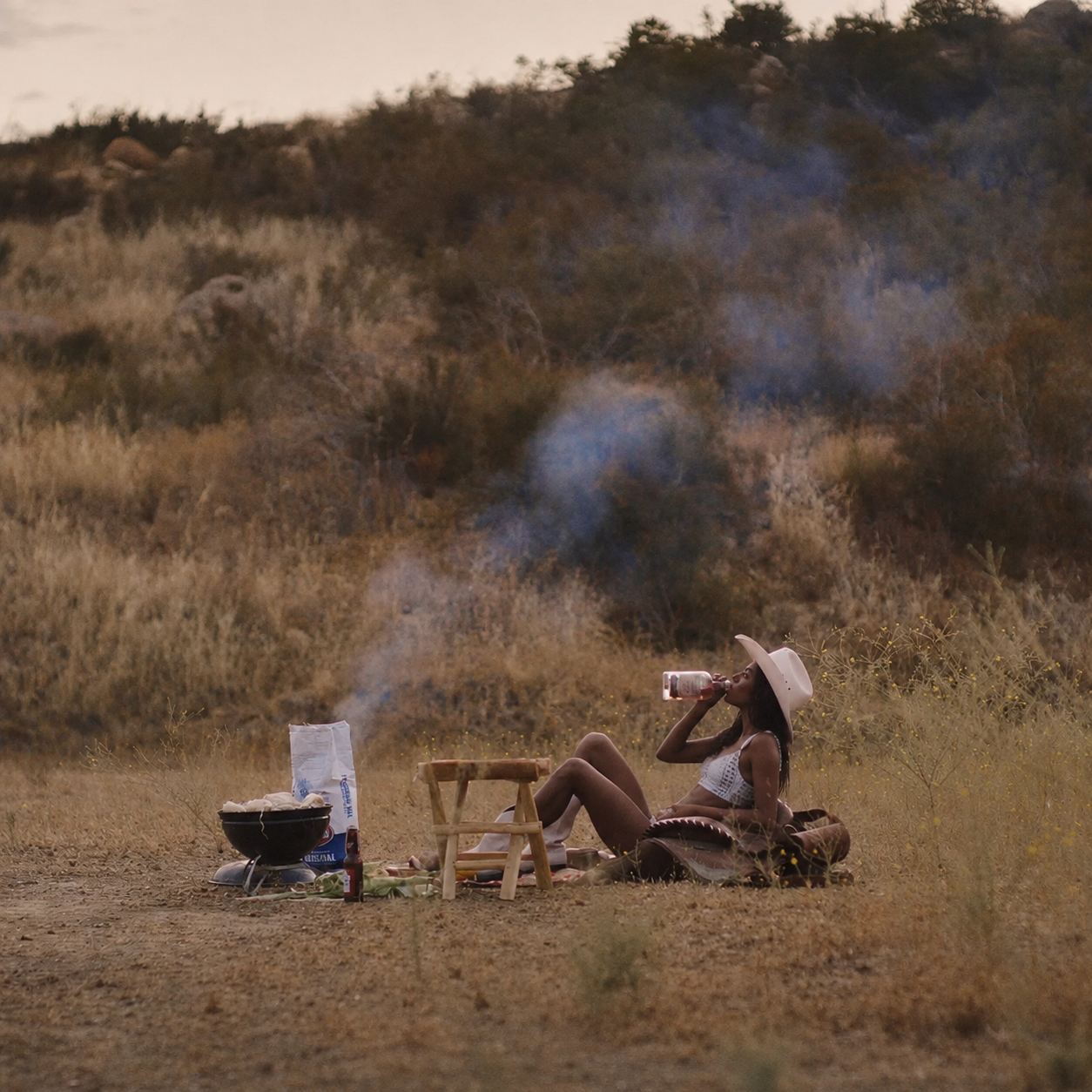 Woman wearing a cowboy hat, sitting alone in field with a BBQ grill, bag of charcoal, drinking from a glass bottle.